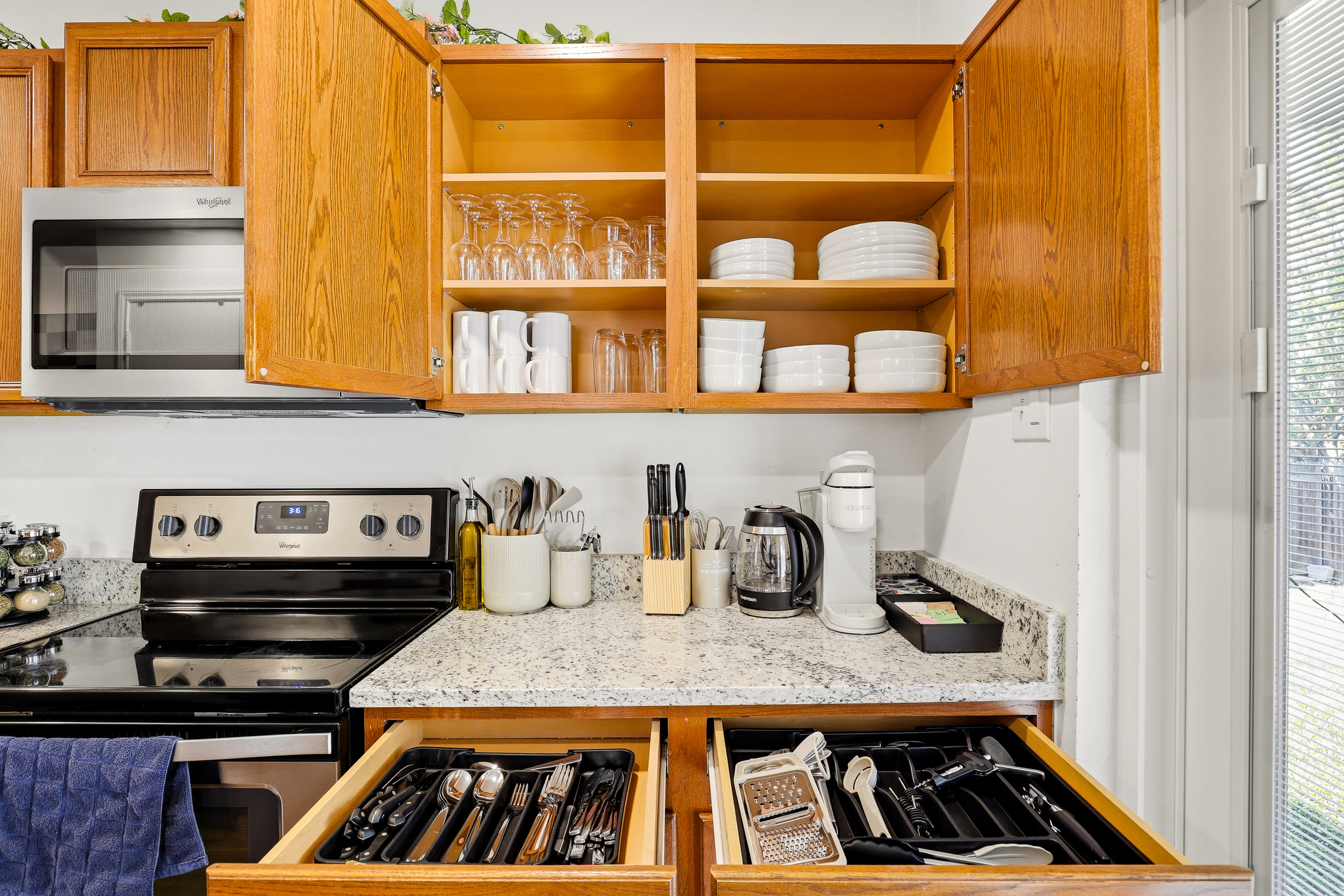 Fully stocked kitchen with wine glasses, plates, mugs, and full silverware drawers — plus electric stove, microwave, and Keurig coffee maker. No supply runs needed, just cook and enjoy.