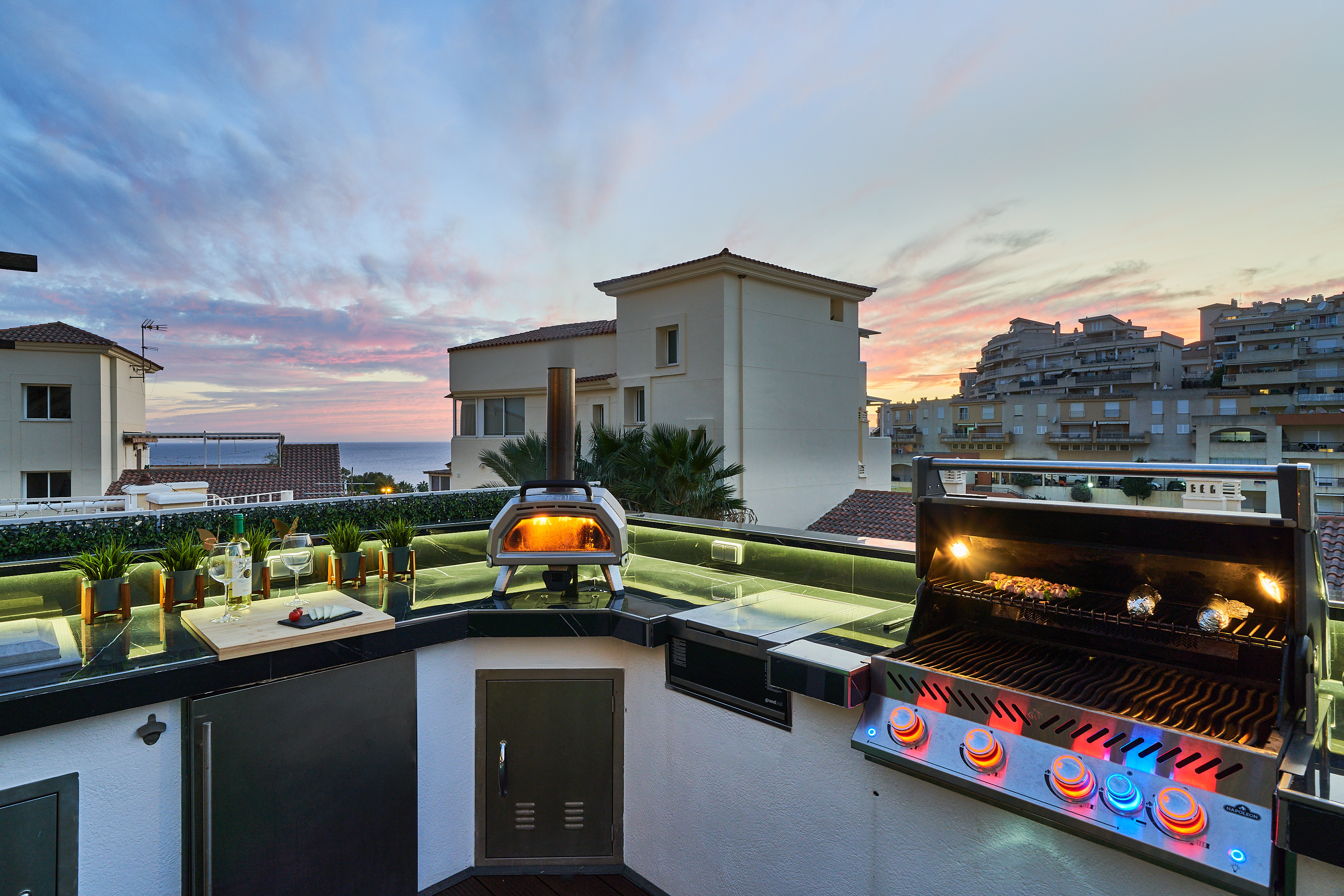 Fire up the built-in barbecue or the wood-fired pizza oven and enjoy cooking with a view. The fully equipped outdoor kitchen features a sink, fridge, and prep space with subtle lighting for evening meals. Dine under the open sky at the outdoor table, surro