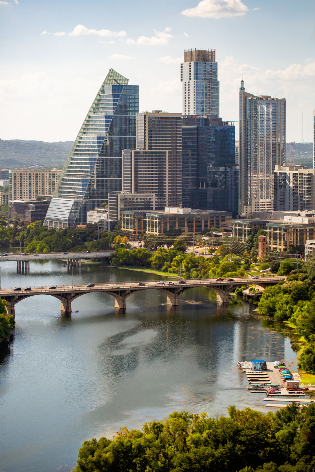 A panoramic view of a vibrant cityscape, showcasing a river with multiple bridges and lush green trees lining its banks. In the background, a stunning skyline of modern high-rise buildings and corporate offices dominates the view, suggesting a bustling urb