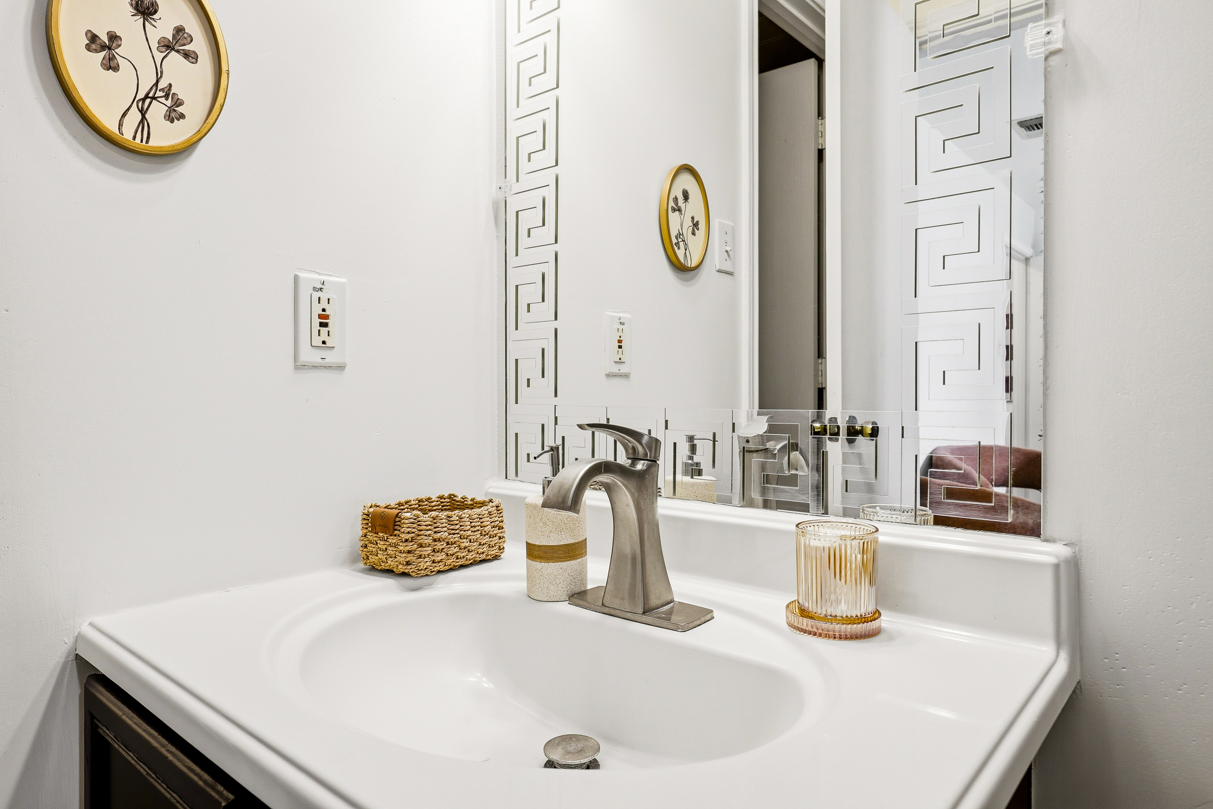 Bathroom vanity detail with framed decorative mirror, woven basket, and brushed nickel faucet. Clean, well-appointed, and stocked with hand soap and cotton swabs for every guest.