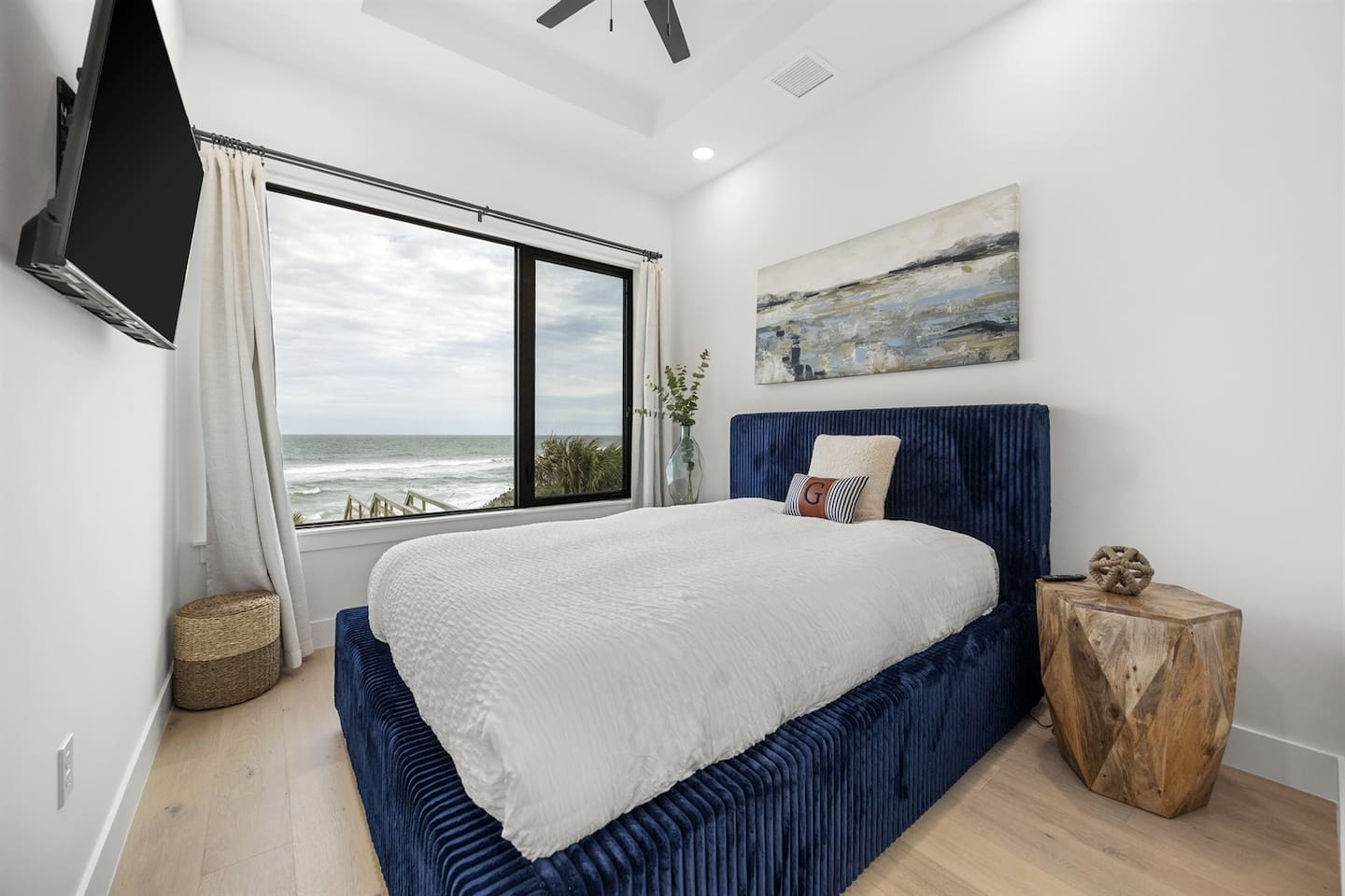 This cozy upstairs bedroom features a queen bed with a stylish dark blue frame and large windows that fill the space with natural light while showcasing stunning ocean views for a peaceful escape.