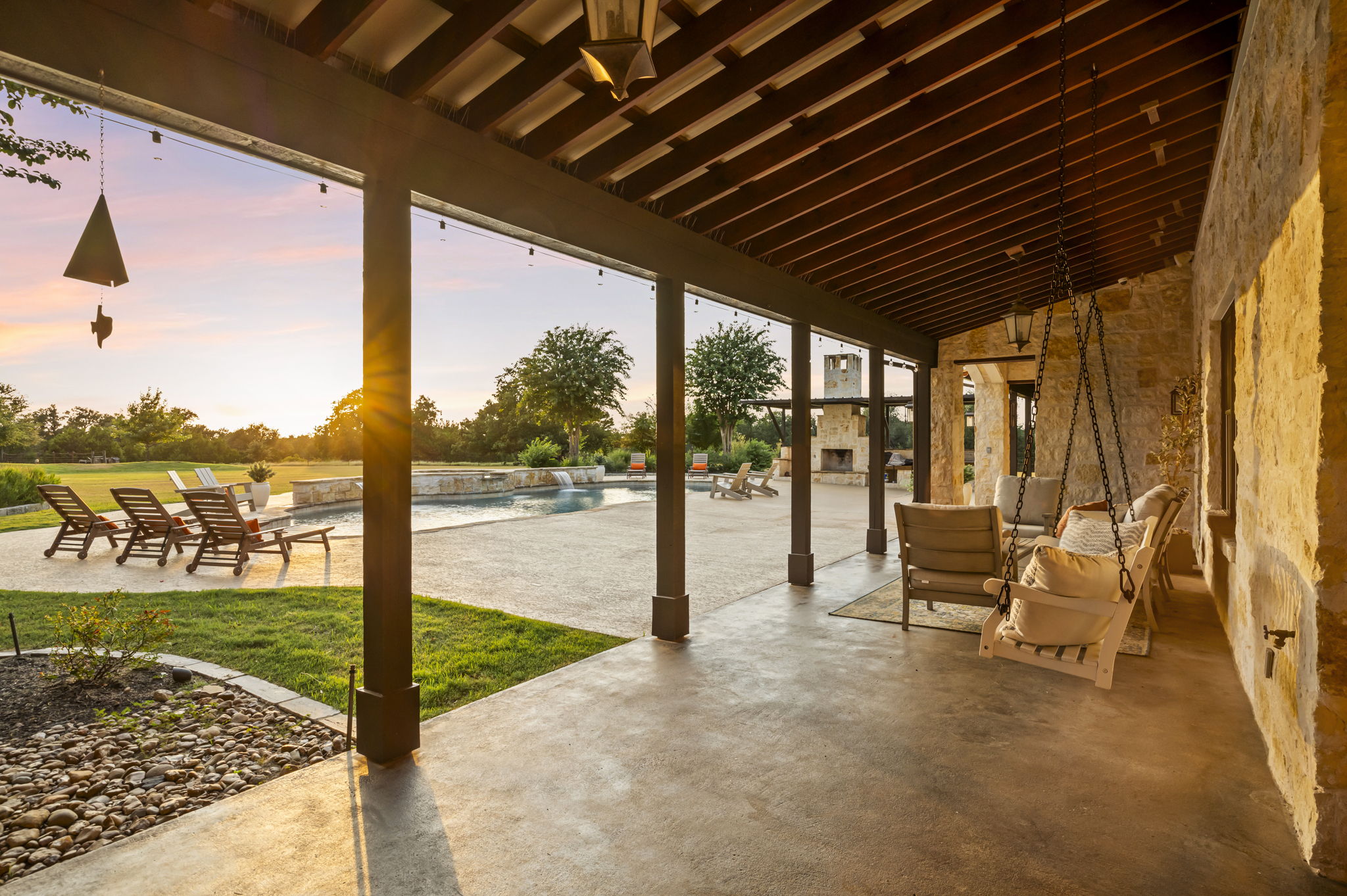 A tranquil evening on a spacious covered patio overlooking a sparkling pool and a lush green lawn, bathed in the warm glow of a setting sun.