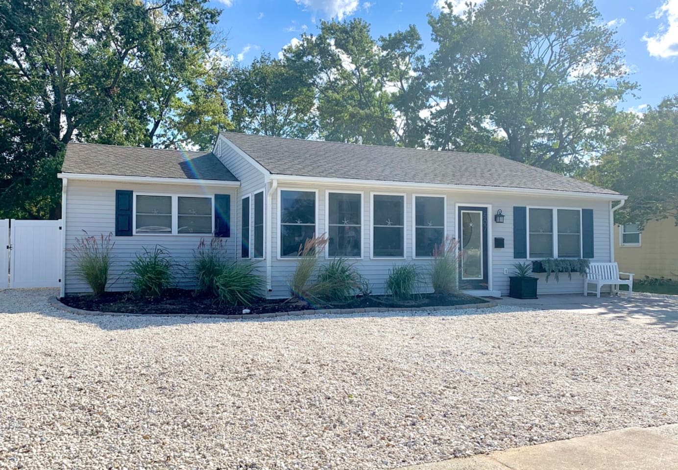 Welcome to your getaway! This lovely ranch-style home features light gray siding, navy shutters, and a bright sunroom. The exterior is complete with a white stone and manicured greenery, offering great curb appeal and a peaceful setting.