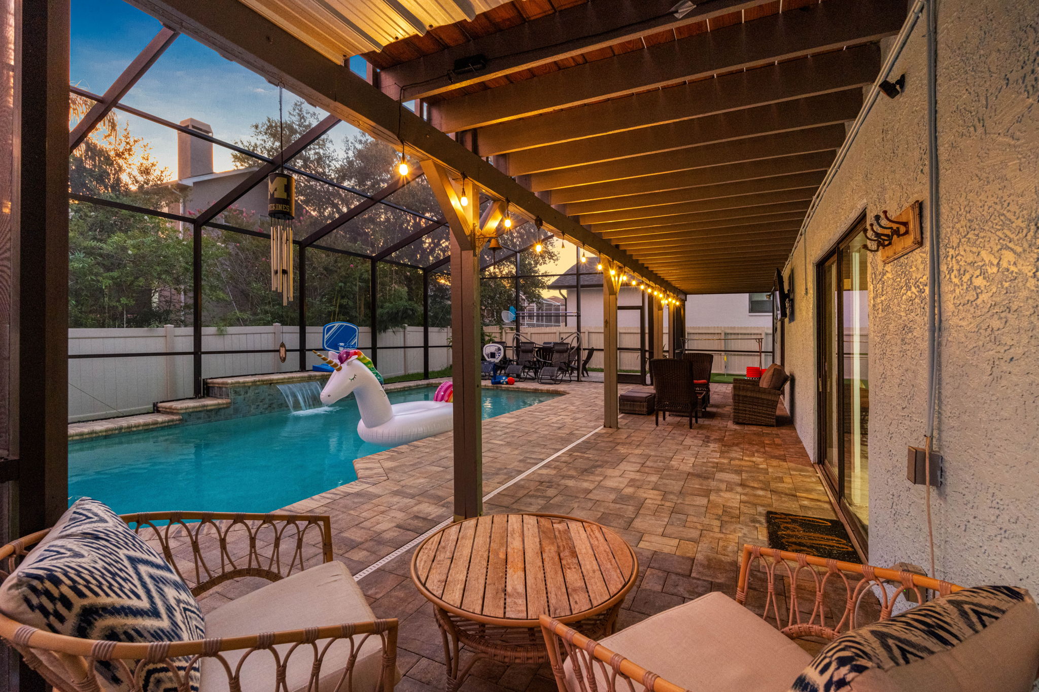 This fully screened backyard retreat features a sparkling pool, playful floaties, cozy conversation areas, and string lights that make evenings feel magical.