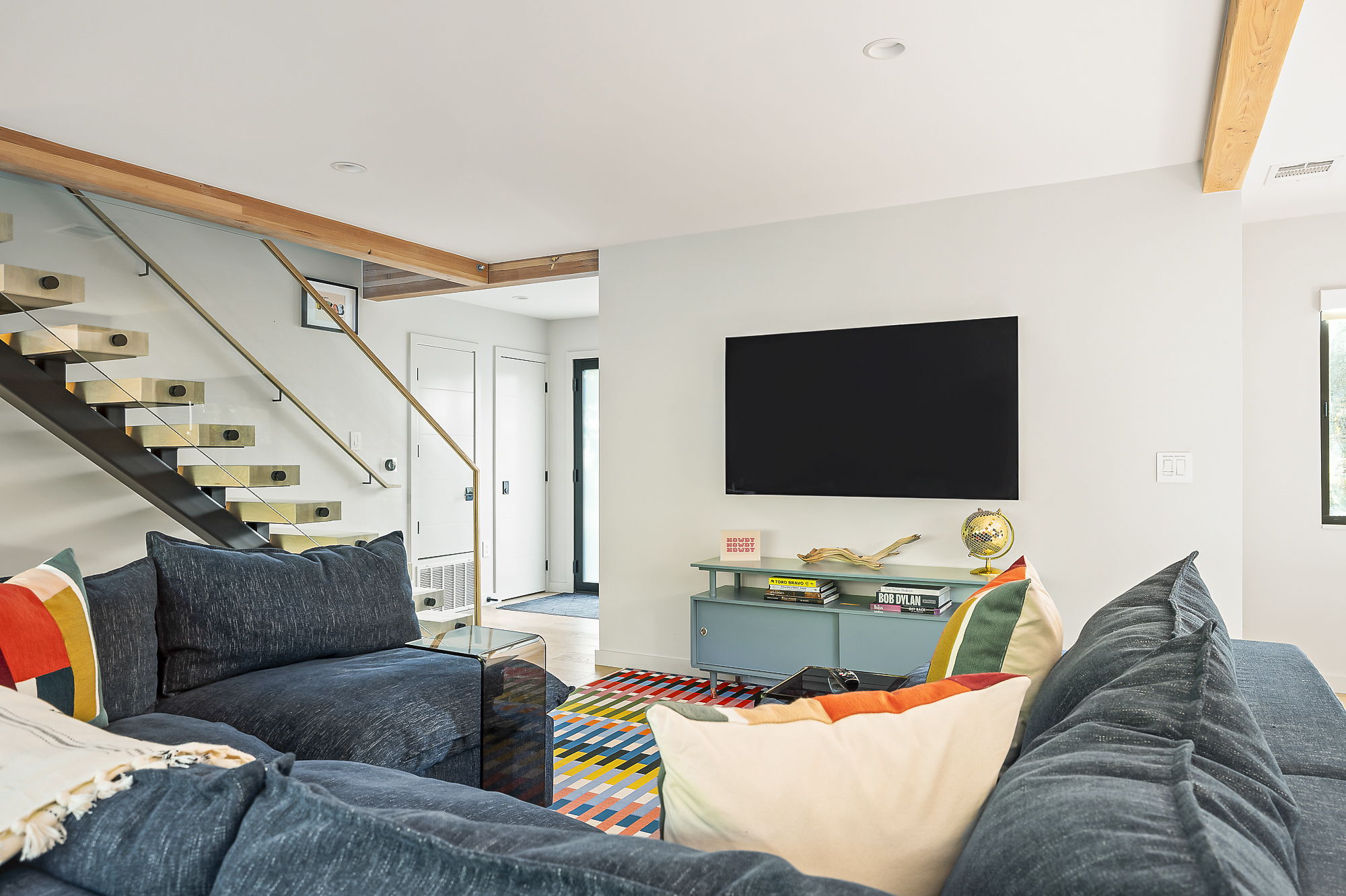 A stylish and vibrant living area with a large sectional sofa, a wall-mounted television above a colorful console, and a modern floating staircase with a glass railing.