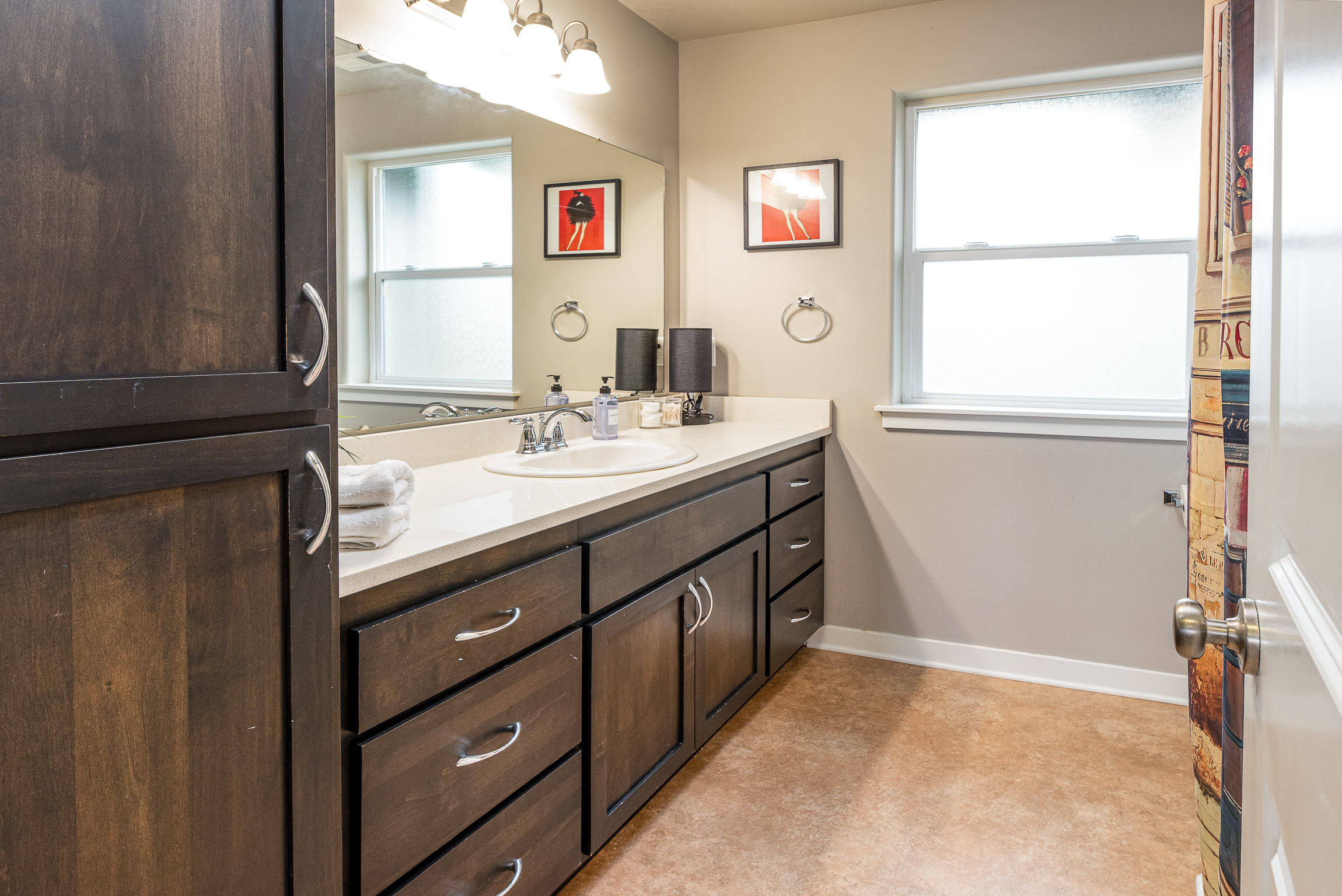 A modern bathroom featuring a double-sink vanity with a light countertop, dark wooden cabinets, a large mirror reflecting the room, two framed artworks on the wall, and a window allowing natural light, with a towel neatly placed on the counter.