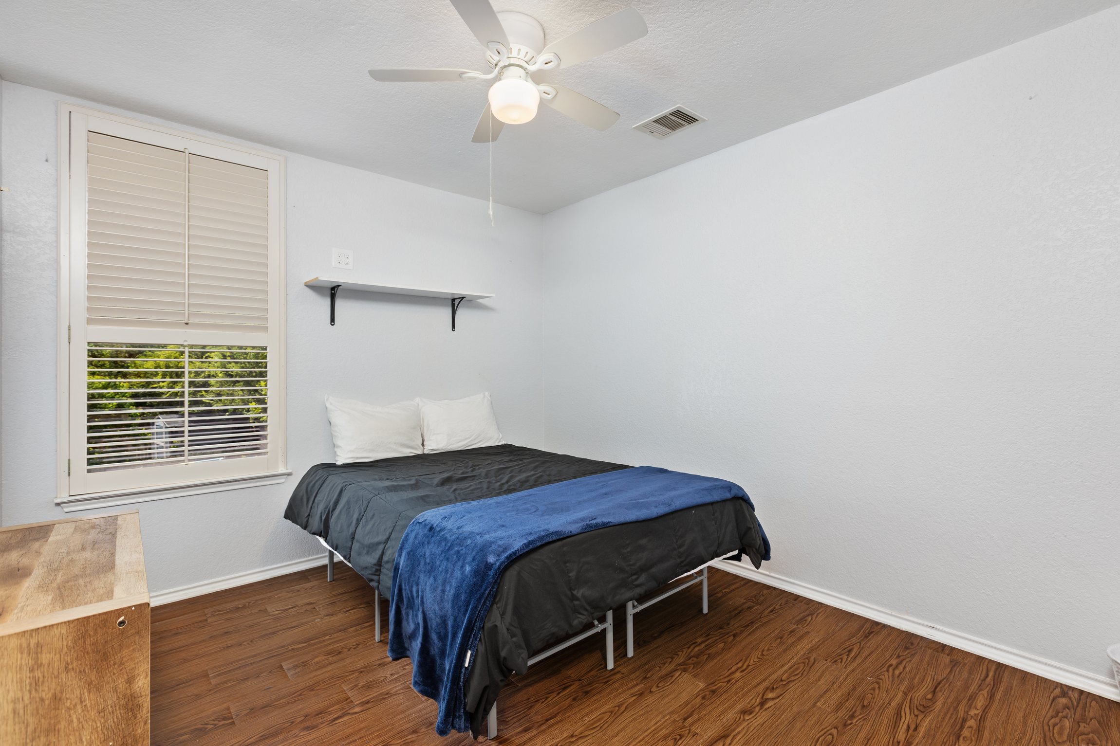 Queen bedroom with soft navy bedding, ceiling fan, window blinds, and clothing storage. Hotel-quality linens and extra pillows included. Quiet and private with hardwood floors in this 5BR San Antonio home.