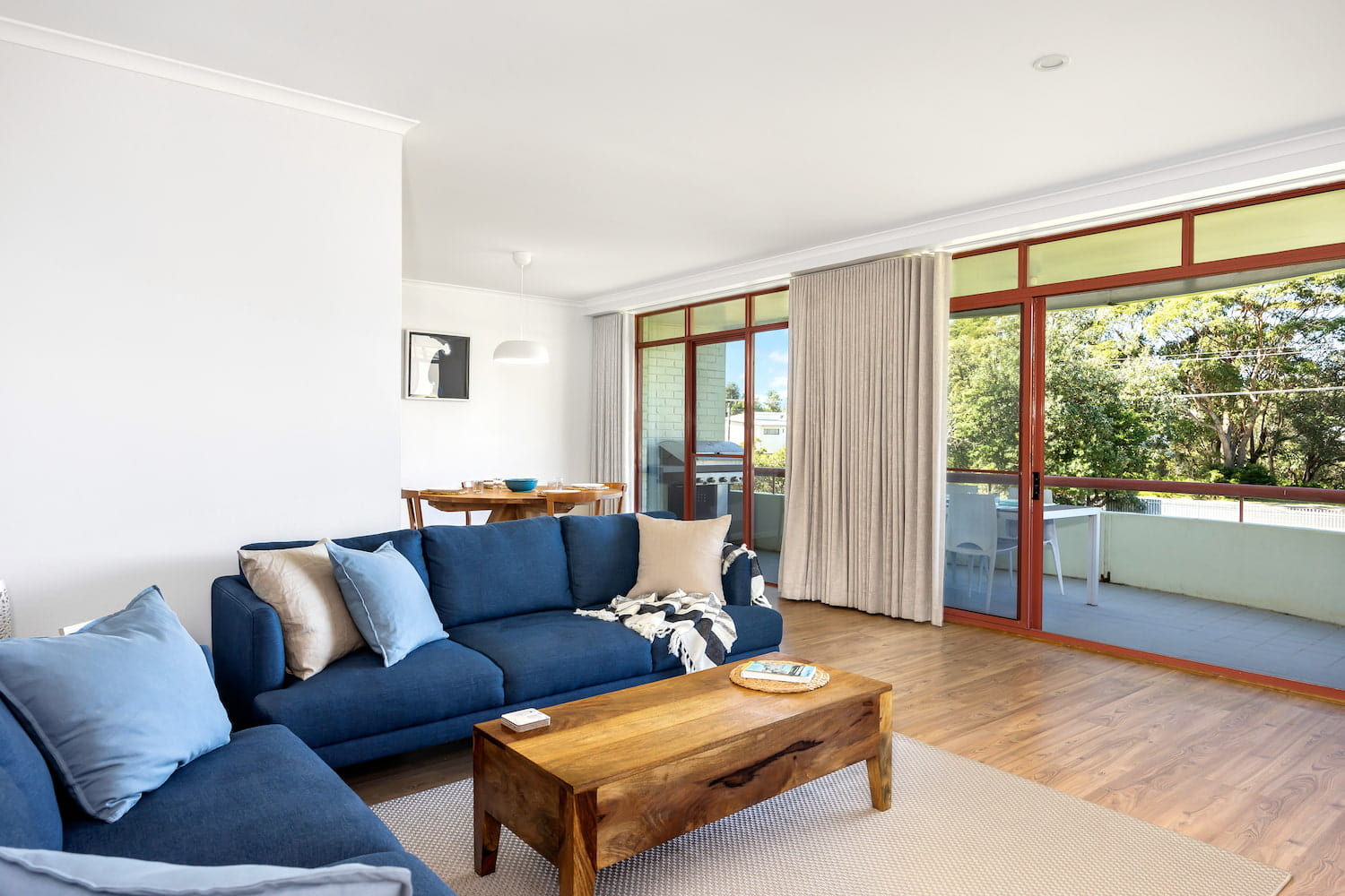 Two blue couches arranged  in L shape plus cushions and wooden coffee table in open plan living area with bottom of dining table visible in the background.