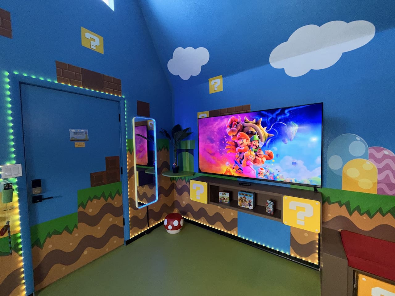 Vibrant blue walls with Super Mario murals, glowing question mark blocks, green pipes, and brick details surround the extra large 75" smart TV (+ Nintendo Switch system). The mirror lights up and plays music via Bluetooth, perfect for family fun!