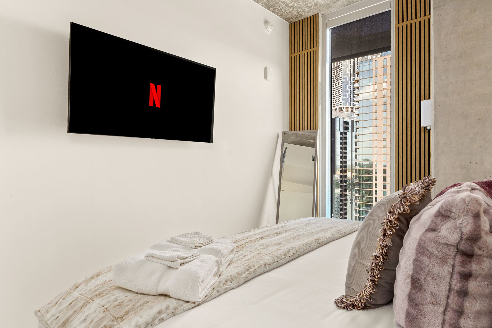 A comfortable bedroom featuring a wall-mounted TV with Netflix on the screen, a large window offering a view of a cityscape, and a full-length mirror, creating a cozy yet modern and stylish retreat.