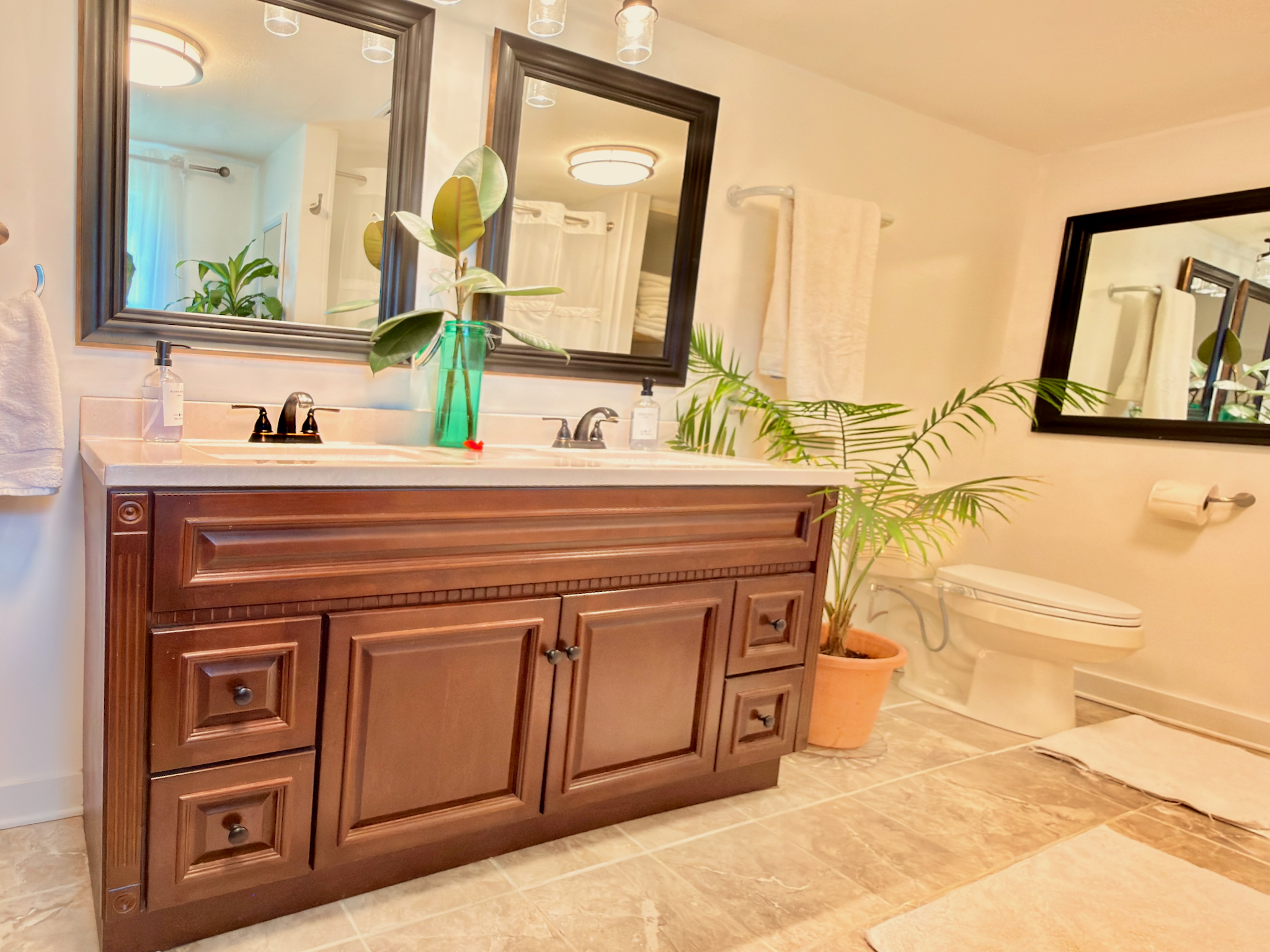 Spa bath with double vanity, bidet commode, tile floor, and organic cotton towels. Features a shower-tub combo (visible in mirror) and living plants.