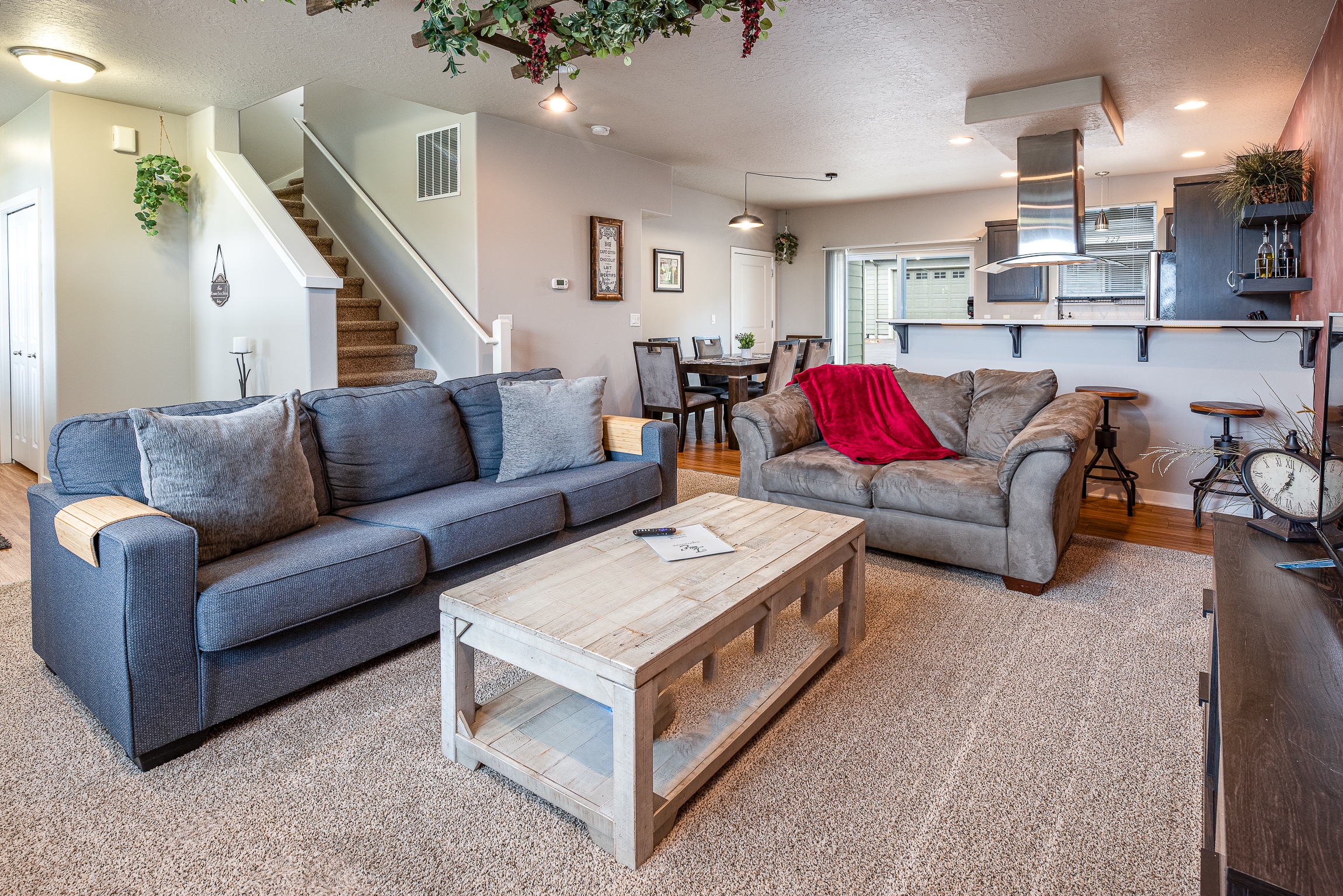 A cozy living room featuring a blue sofa, a gray loveseat with a red throw blanket, a rustic wooden coffee table, and a dining area with a dark wooden table and chairs, along with a staircase leading to the upper floor, all set against a neutral-colored wall adorned with greenery and modern lighting.
