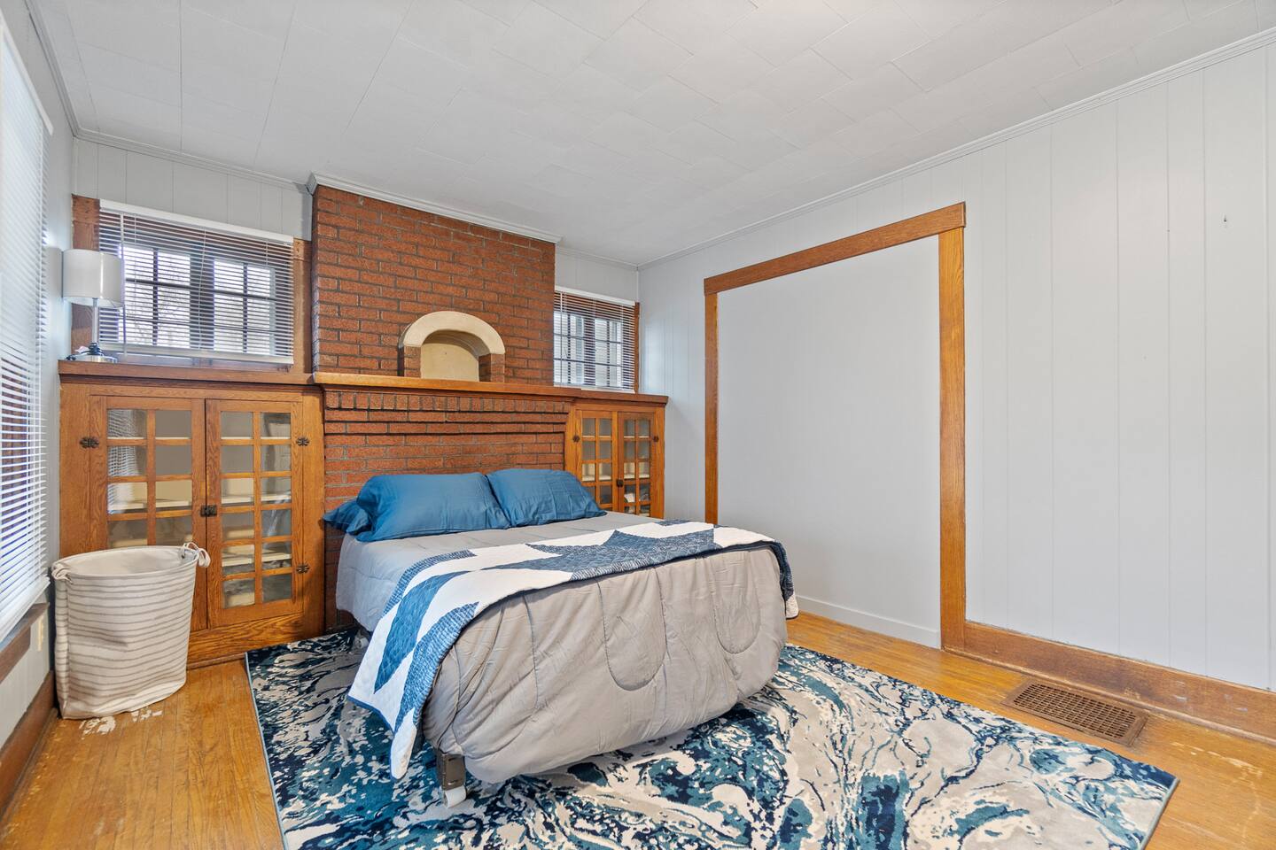 Cozy bedroom featuring a modern sofa with pillows, large windows next to the bed, a sleek wood cabinet, a stylishly designed backdrop behind the bed, all set against a crisp white wall—creating a comfortable and inviting space with contemporary charm