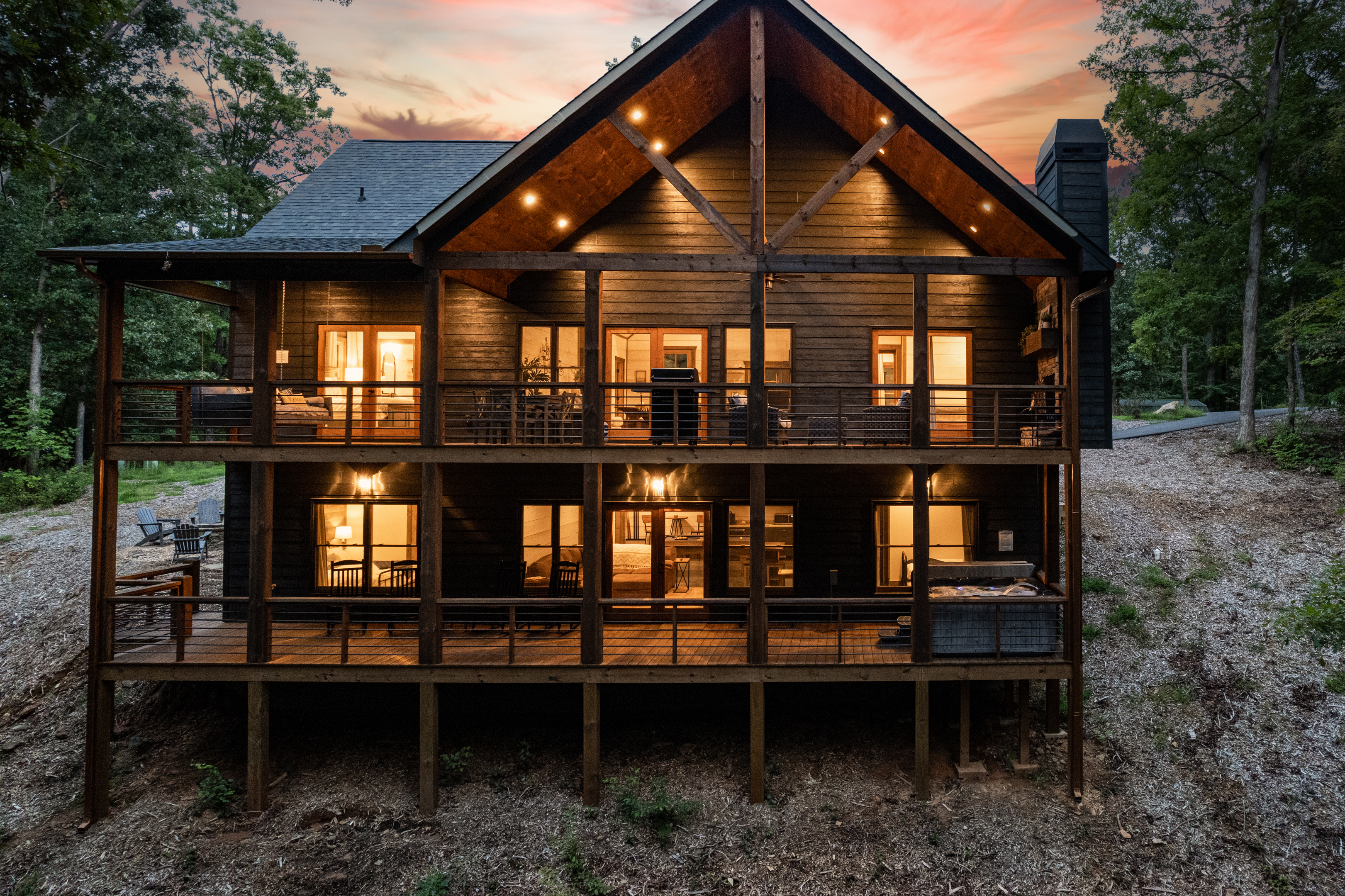 Rear exterior of the cabin showing layout of decks with hot tub, and rockers on lower level and fireplace, grill, eating and sitting areas and hanging swing on upper level.