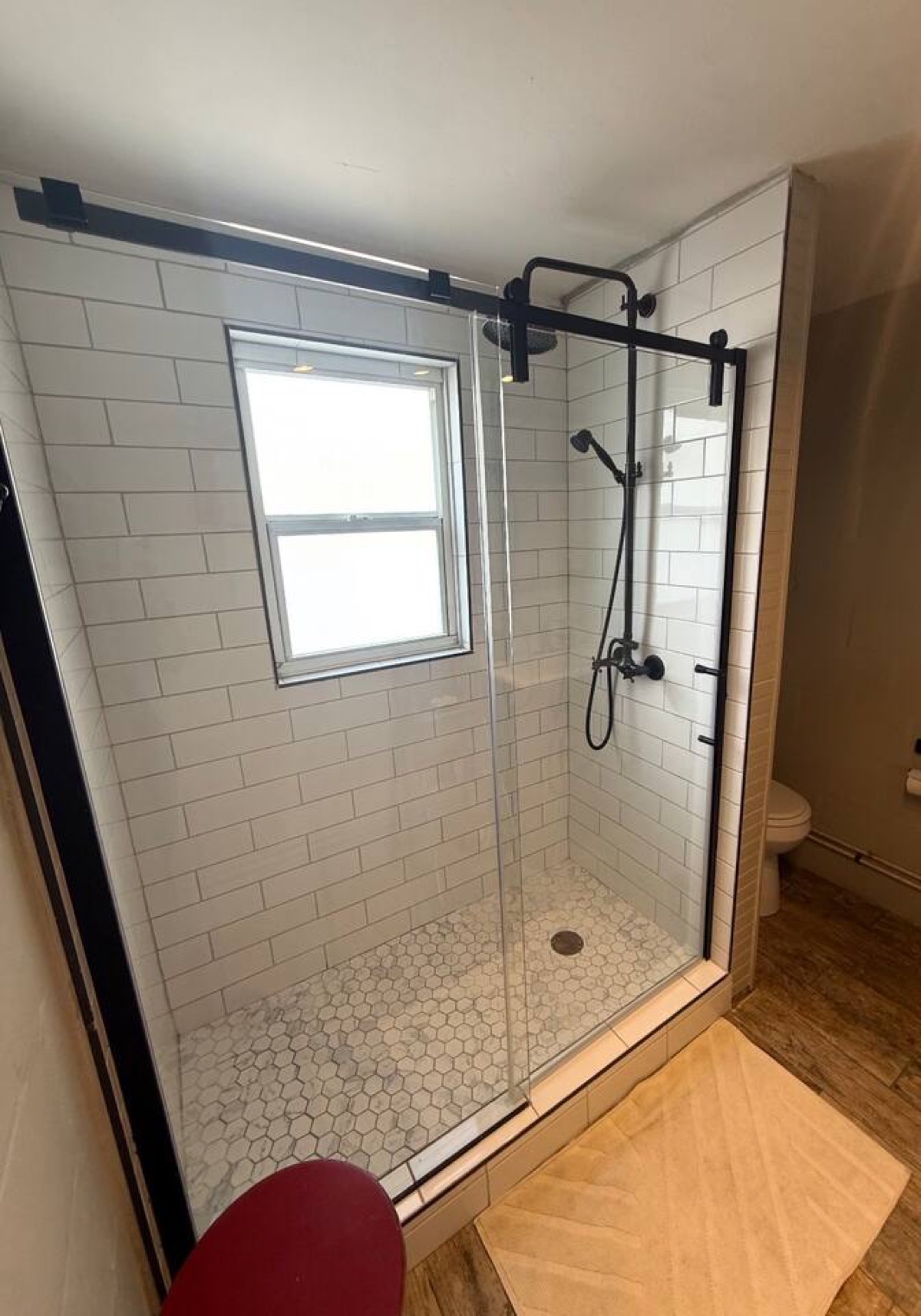 Newly upgraded bright, modern walk-in shower w/ white subway tile and classic hex tile floor. Features black rain shower head, handheld sprayer, and sliding glass barn-style door. Natural light from the frosted window keeps the space airy and private