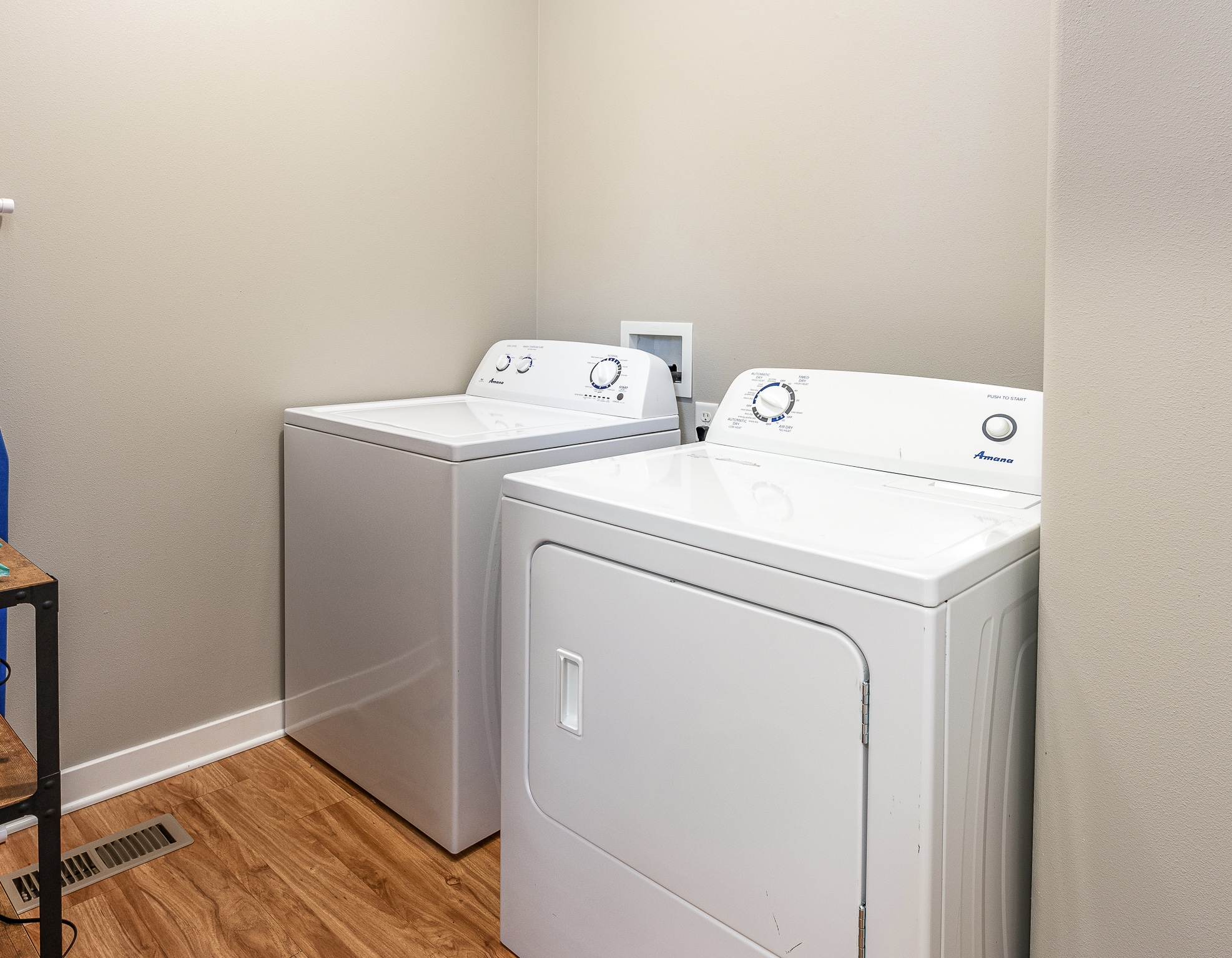 A laundry room features a white top-loading washing machine and a white front-loading dryer positioned side by side against a light beige wall, with a wooden shelf and floor vent visible in the foreground.