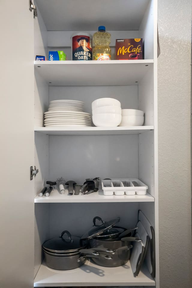 View of a well-organized cabinet stocked with dinnerware, silverware, pots and pans, and cutting boards, providing all the essentials for cooking and dining.