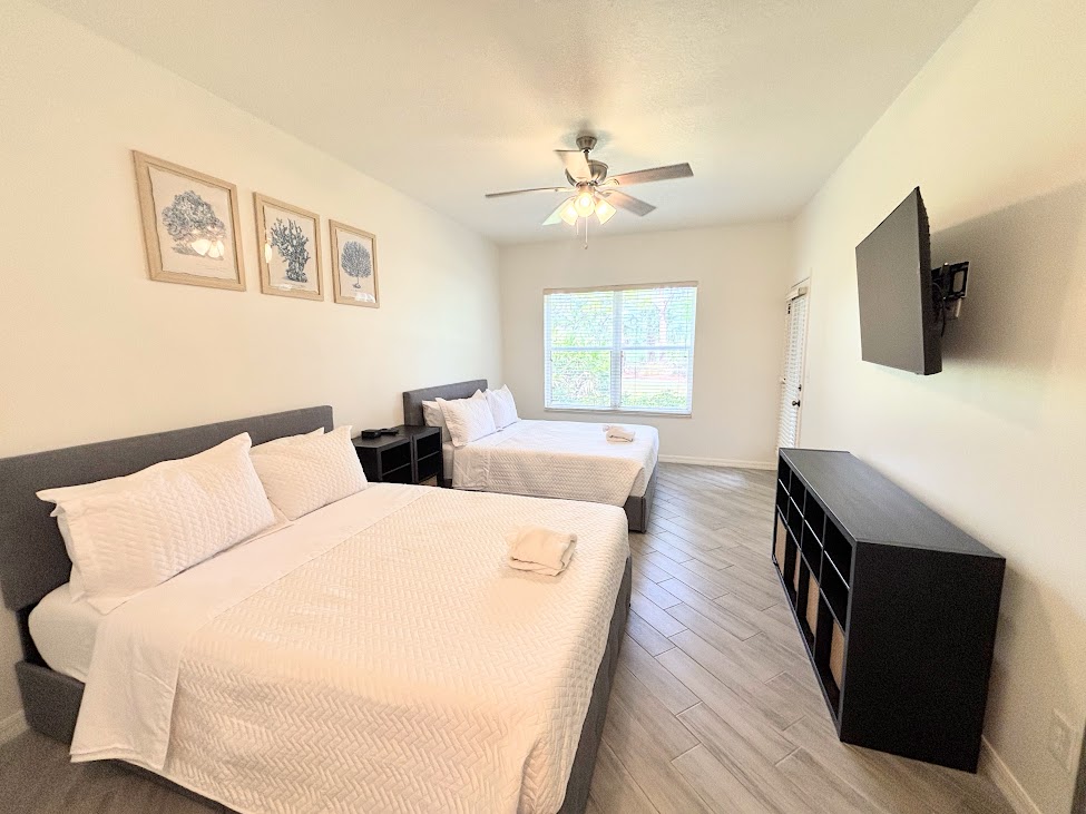 Spacious bedroom featuring two beds, wall-mounted TV, dresser storage, and wood-look flooring.