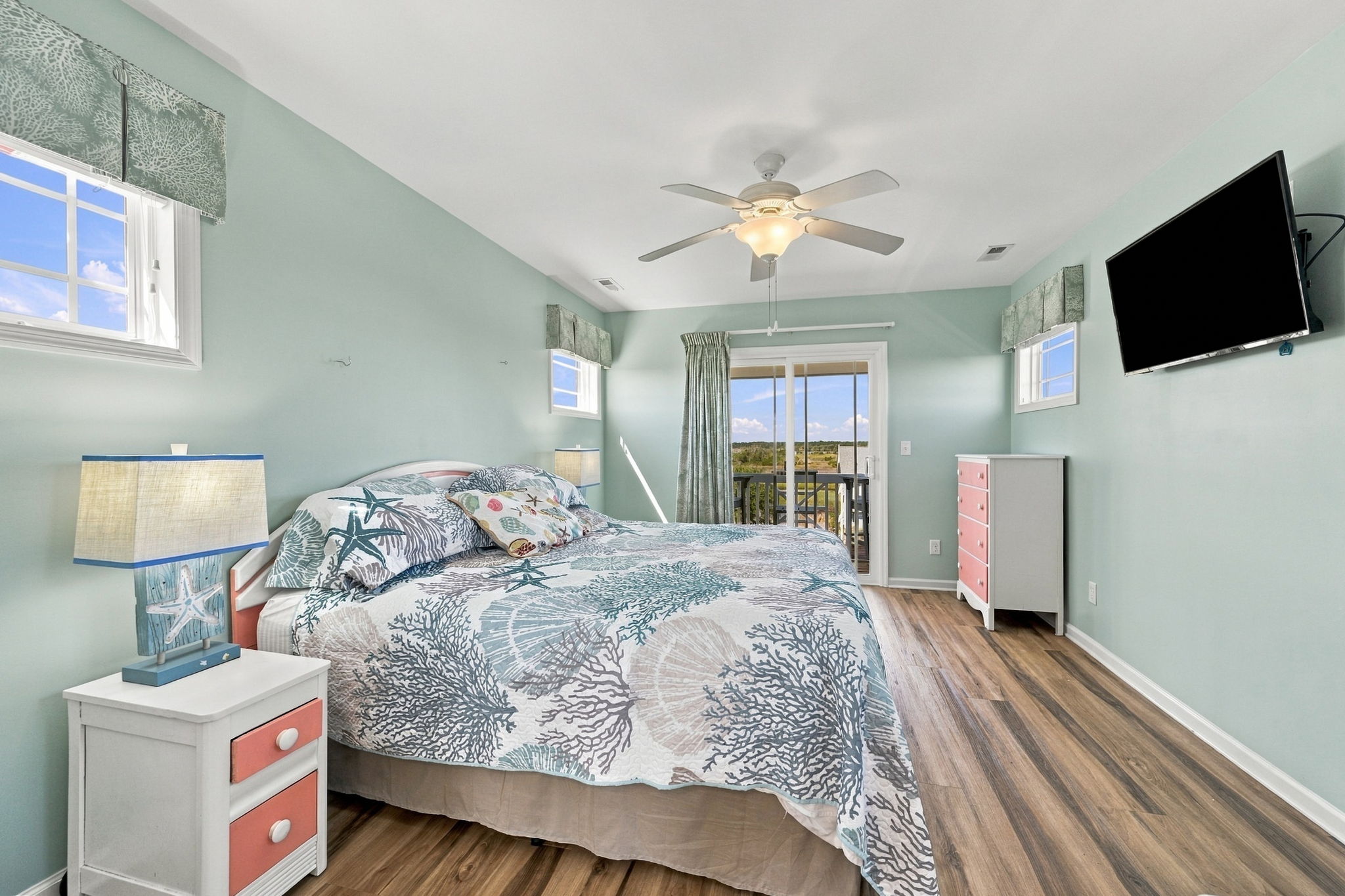 Comfortable coastal-inspired bedroom featuring a spacious bed, wall-mounted TV, and plenty of natural light