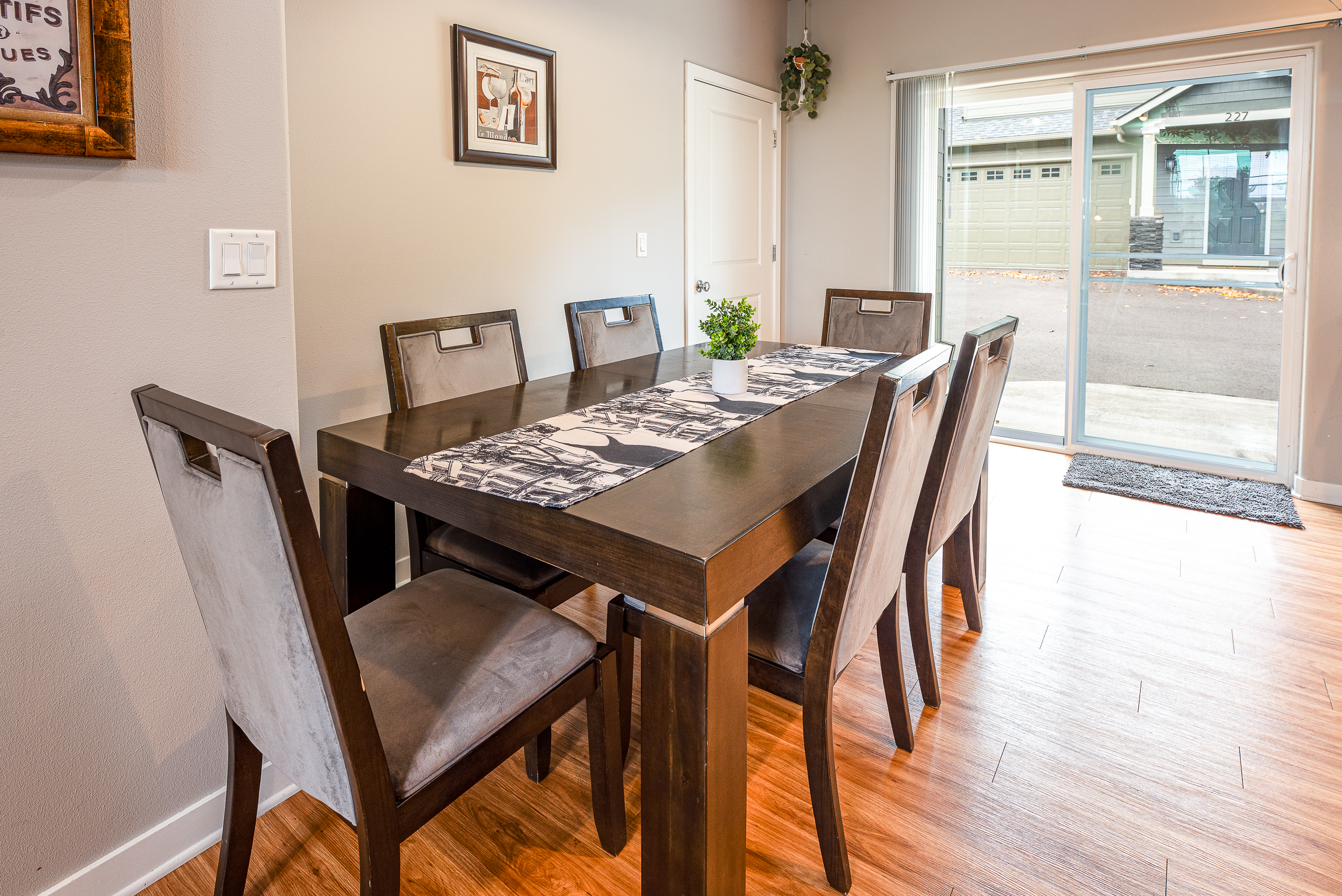 A modern dining area featuring a dark wooden table surrounded by six upholstered chairs, adorned with a black and white table runner and a small potted plant, with a sliding glass door revealing an outdoor view.