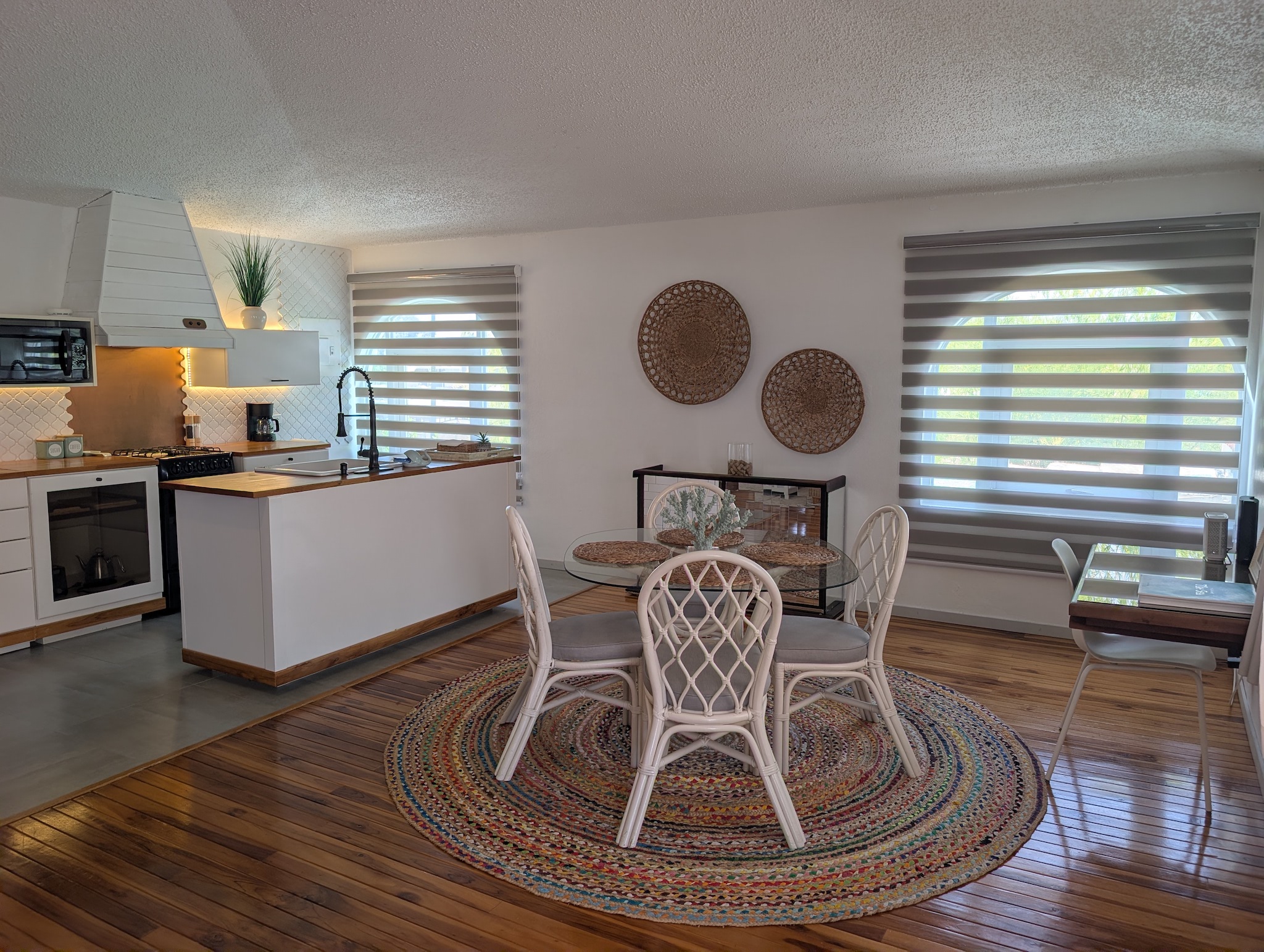 Open-concept upstairs kitchenette and dining area at Villa Coral featuring a round glass table with white chairs, a woven jute rug, hardwood floors, and modern white cabinetry in Dorado, Puerto Rico.
