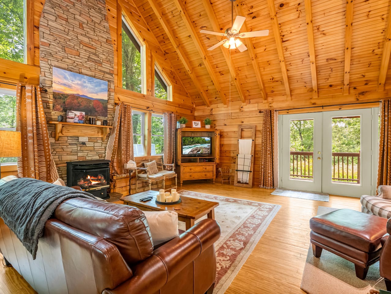 Main living area featuring high vaulted wooden ceilings with exposed beams, stone fireplace with a mantel, french doors opening to the back deck, television set, leather sofa and ottoman, and a wooden coffee table