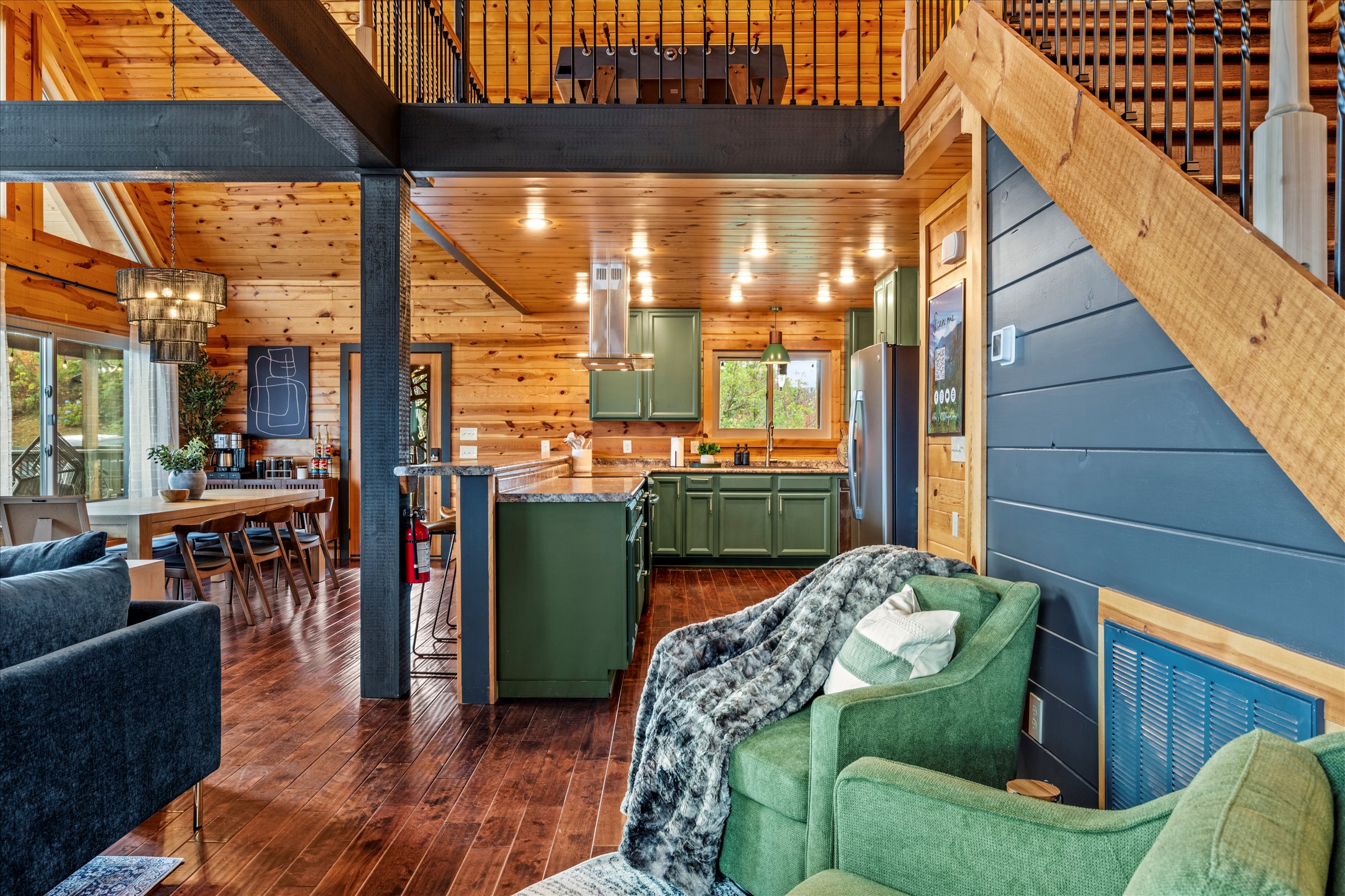 Open-concept living area featuring a modern green kitchen, cozy fireside seating, and a view of the game loft above.