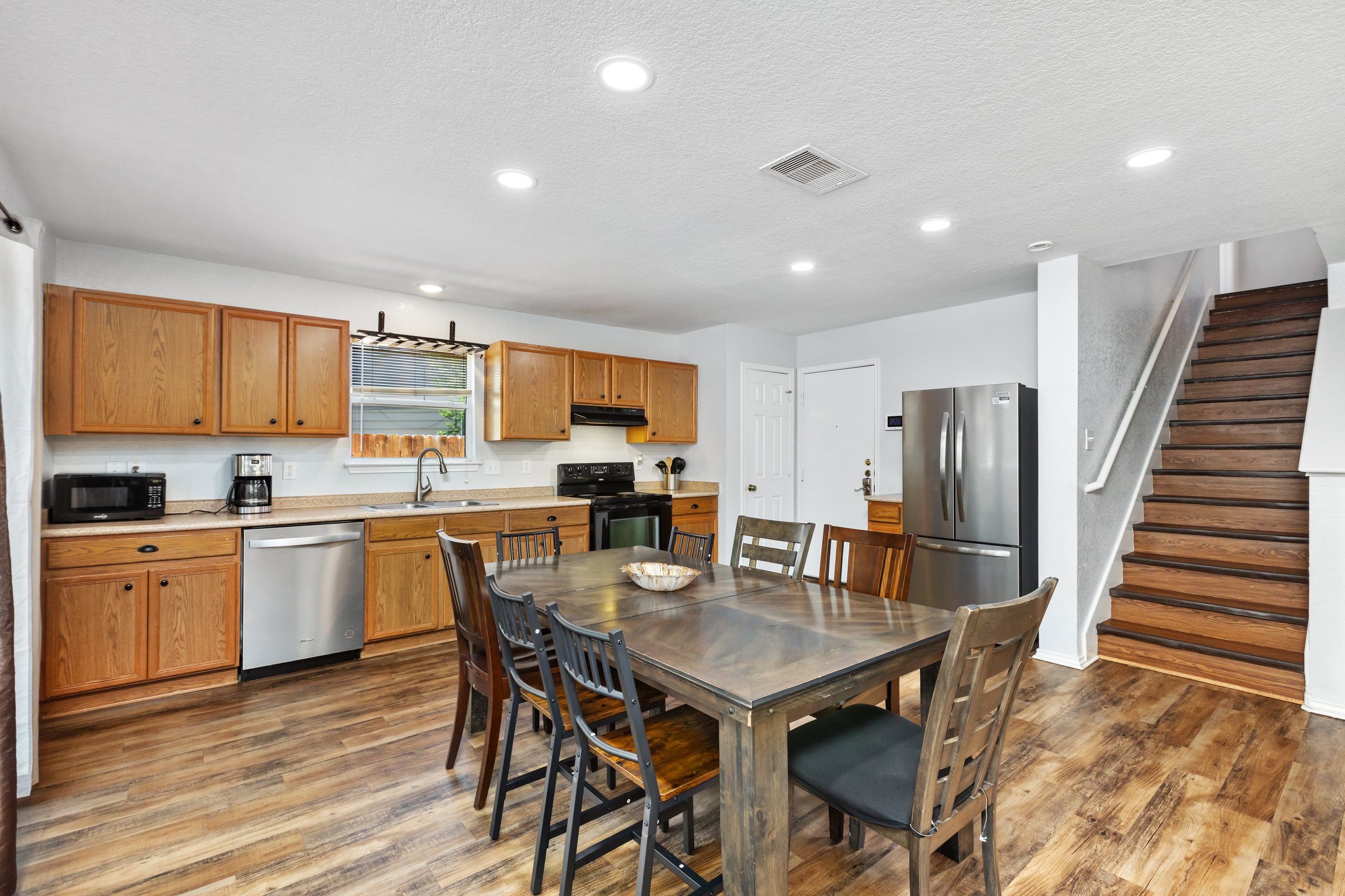Open-concept main floor with kitchen, dining table, stainless refrigerator, and living room all connected. Hardwood floors and staircase leading to 4 upstairs bedrooms. Northeast San Antonio group rental, sleeps 10.