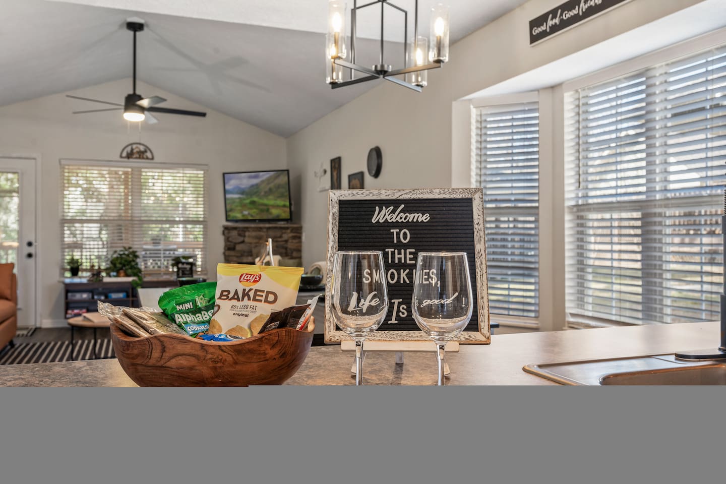 Bright open-concept room with vaulted ceilings, large windows, cozy seating, a stone fireplace, and a welcome station with snacks and wine glasses.