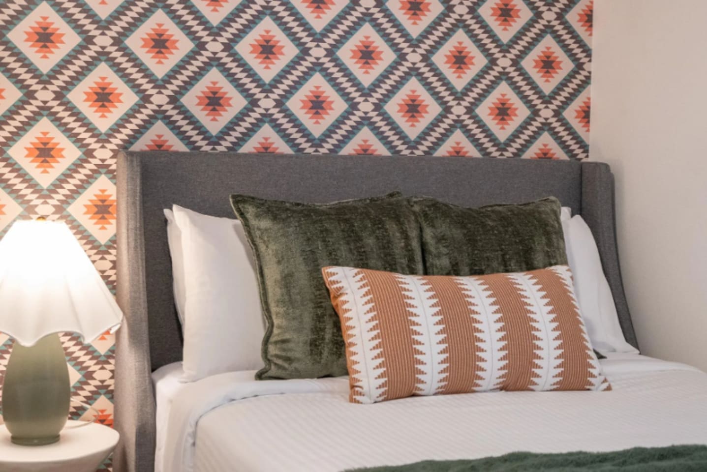 A bold Southwest-print wall backs a plush upholstered headboard, layered with olive velvet and rust-striped pillows for a cozy, textured finish.
