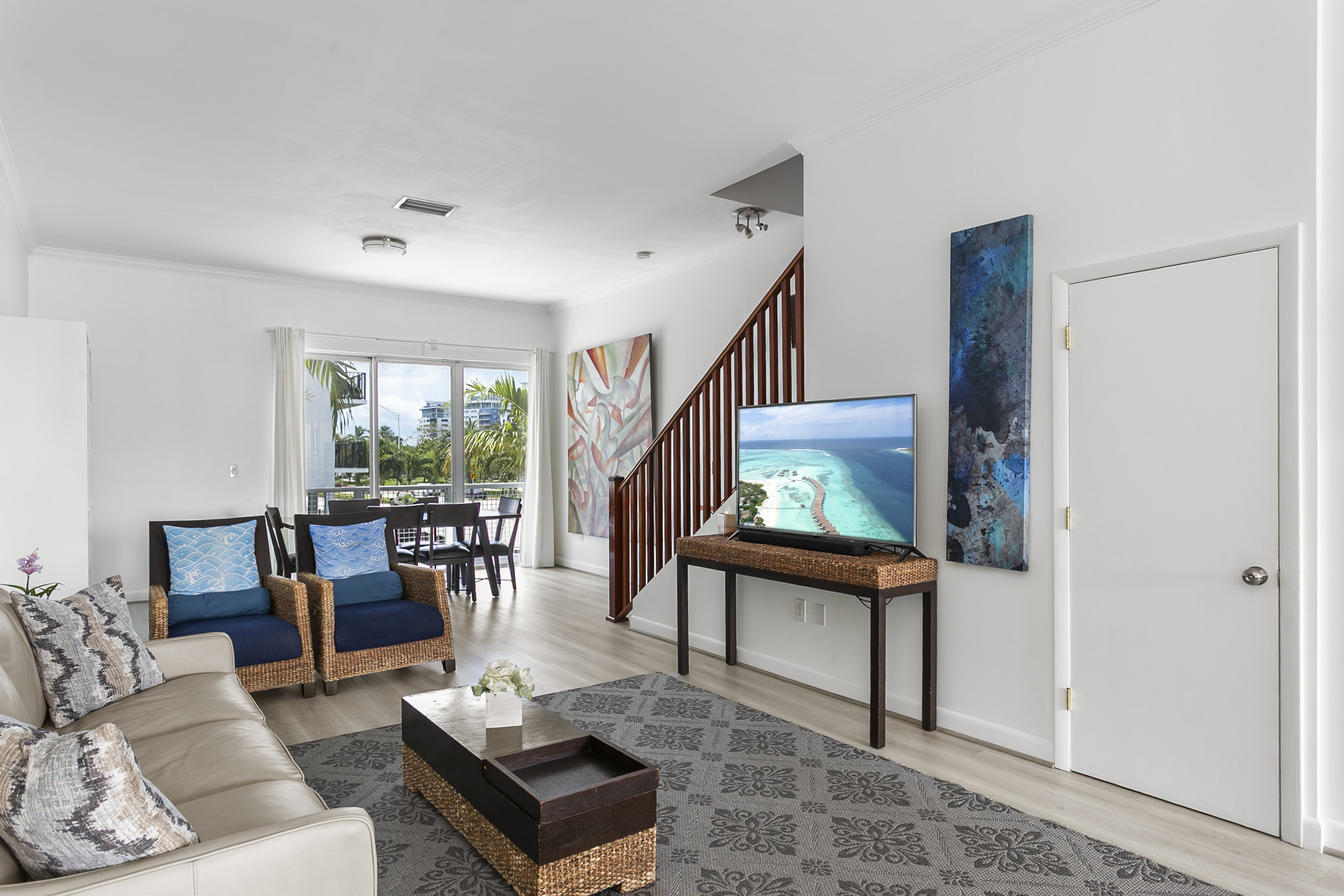 Living room with leather sofa, accent chairs, and a 50-inch flat screen TV. Sliding glass doors open to a Juliet balcony for sea breeze.