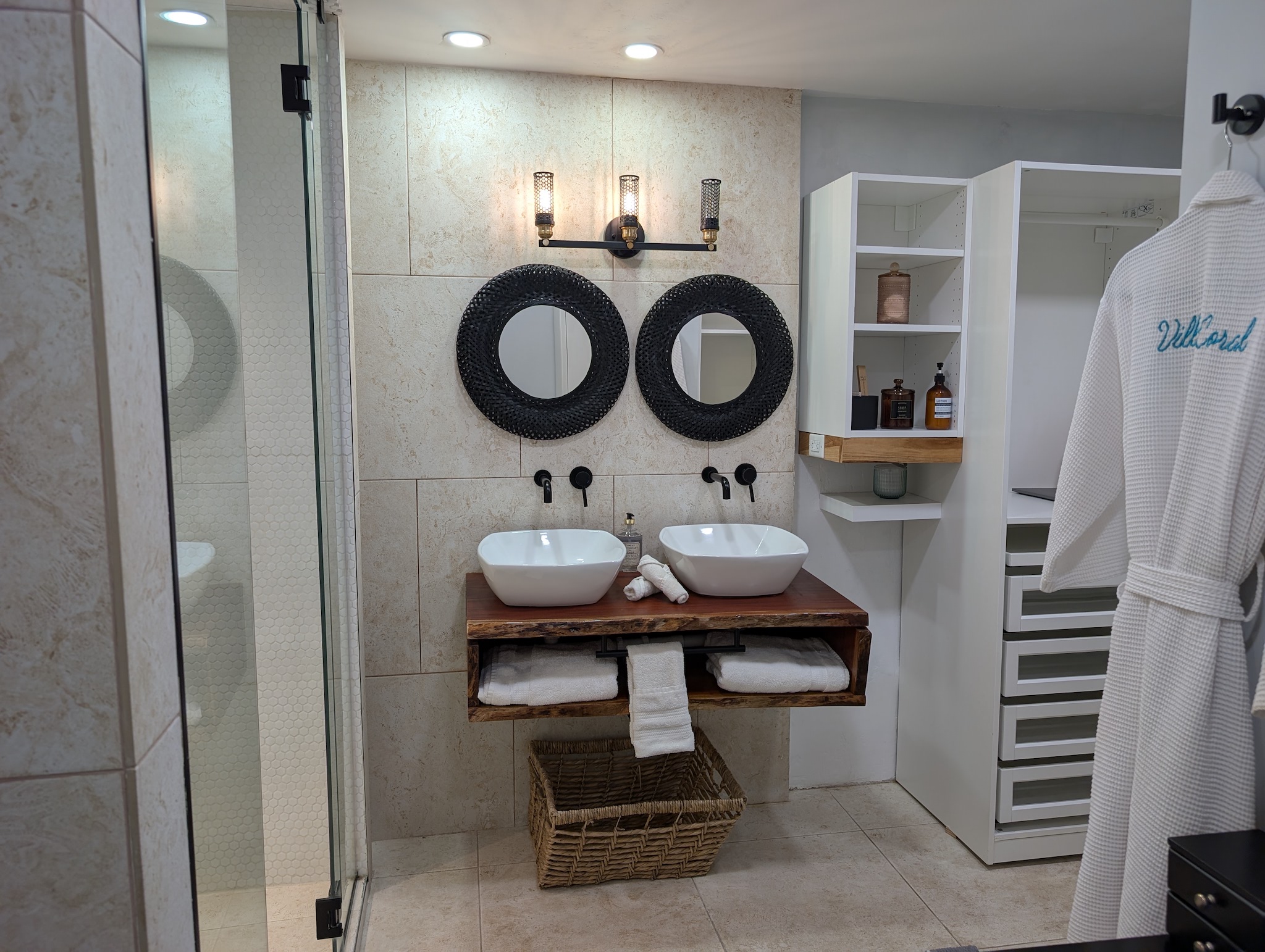 Symmetrical view of the primary bathroom at Villa Coral displaying a custom live-edge wood vanity, vessel sinks, a glass steam shower, built-in closet organization, and travertine tile walls in Dorado, Puerto Rico.
