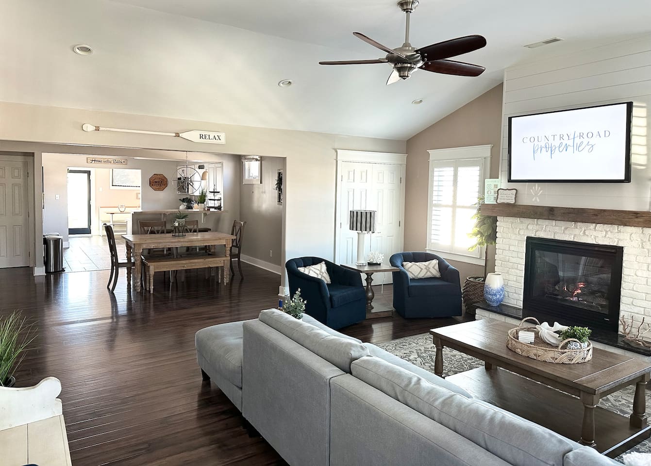 This open-concept layout connects the living and dining areas, featuring vaulted ceilings and a cozy fireplace. The space includes a large sectional and a farmhouse-style dining table, perfect for family meals or a movie night.