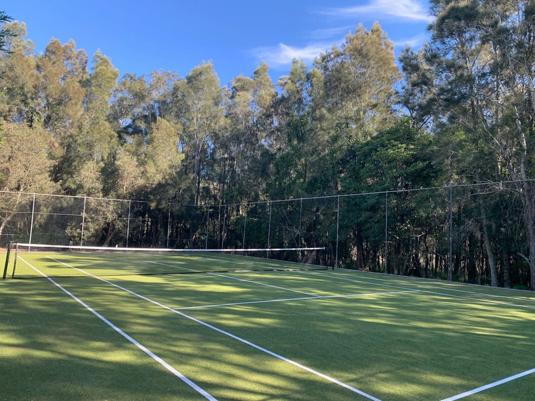 Fathoms tennis court with high tennis court fence, net, astro turf surface and shadows from the surrounding tall trees.
