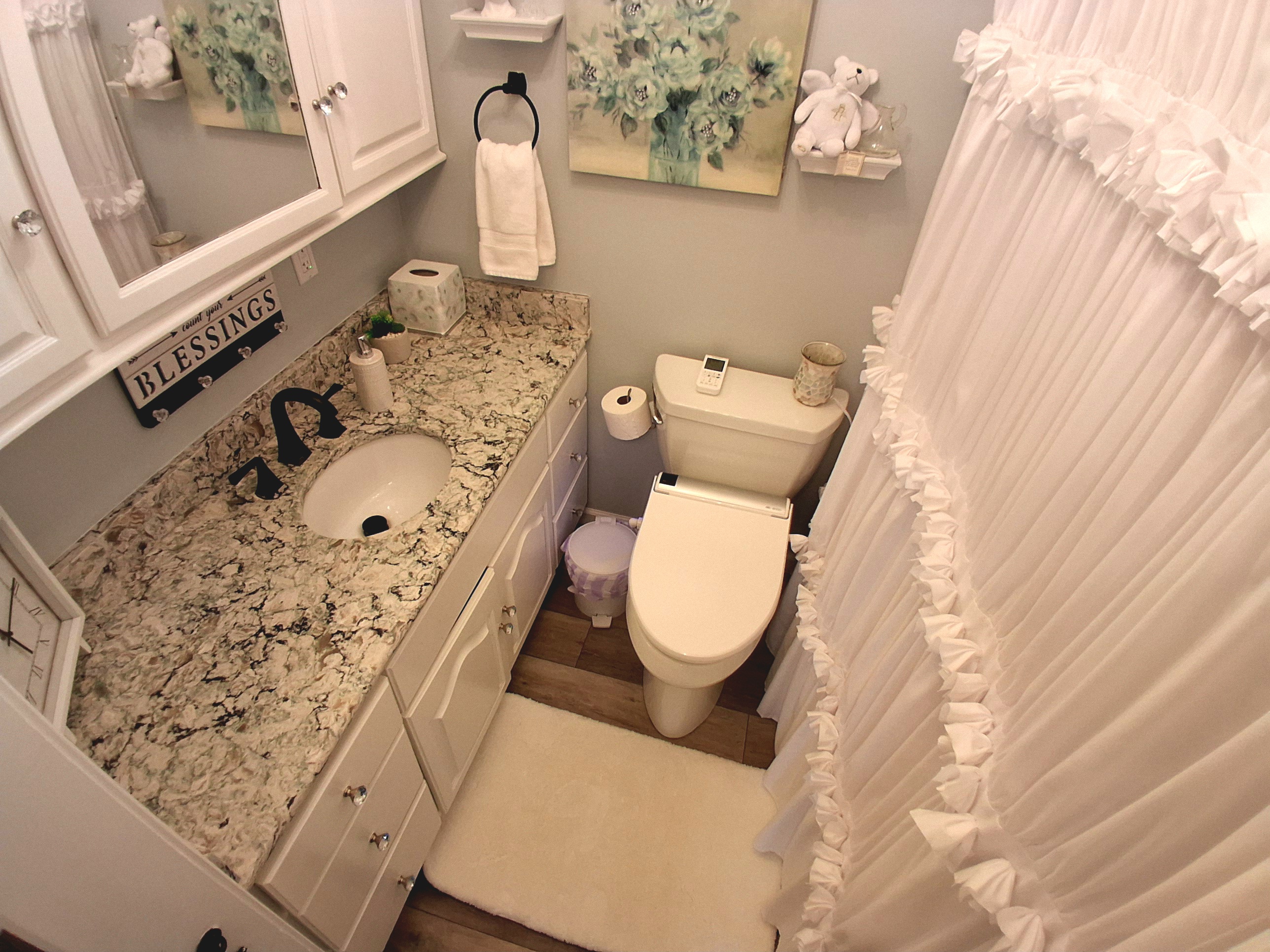 Pale blue walls offer a refined contrast to the crisp white décor, with a romantic ruffled shower curtain and vintage-style vanity. A sparkling marbled countertop adds beauty and space, creating a serene, elegantly styled sanctuary.