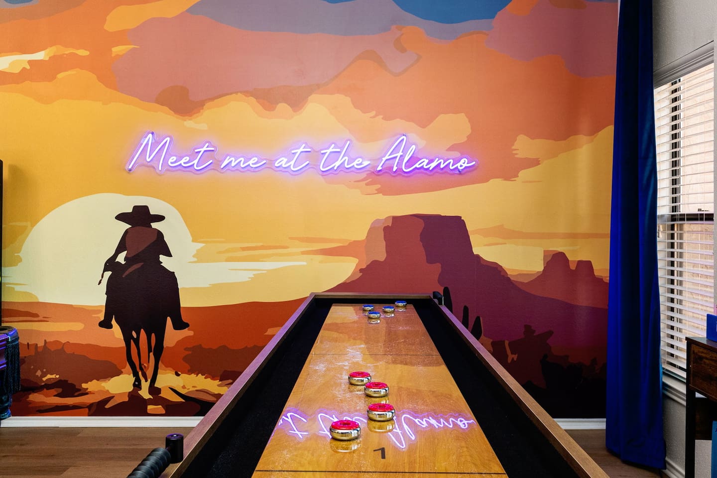 This vibrant Western-themed game room features a shuffleboard table set against a bold mural with a cowboy silhouette and neon “Meet me at the Alamo” sign—bringing Texas pride and playful energy to every game night.