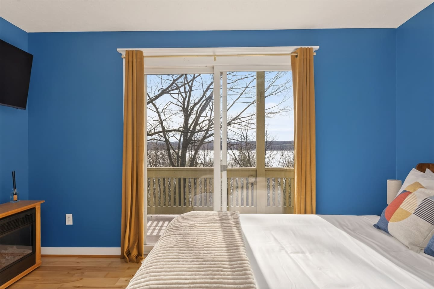 Bright blue master bedroom with stylish artwork, Smart TV, and private balcony access—wake up to peaceful lake views, step outside to sip your morning coffee, and enjoy a calm, relaxing retreat filled with light and comfort