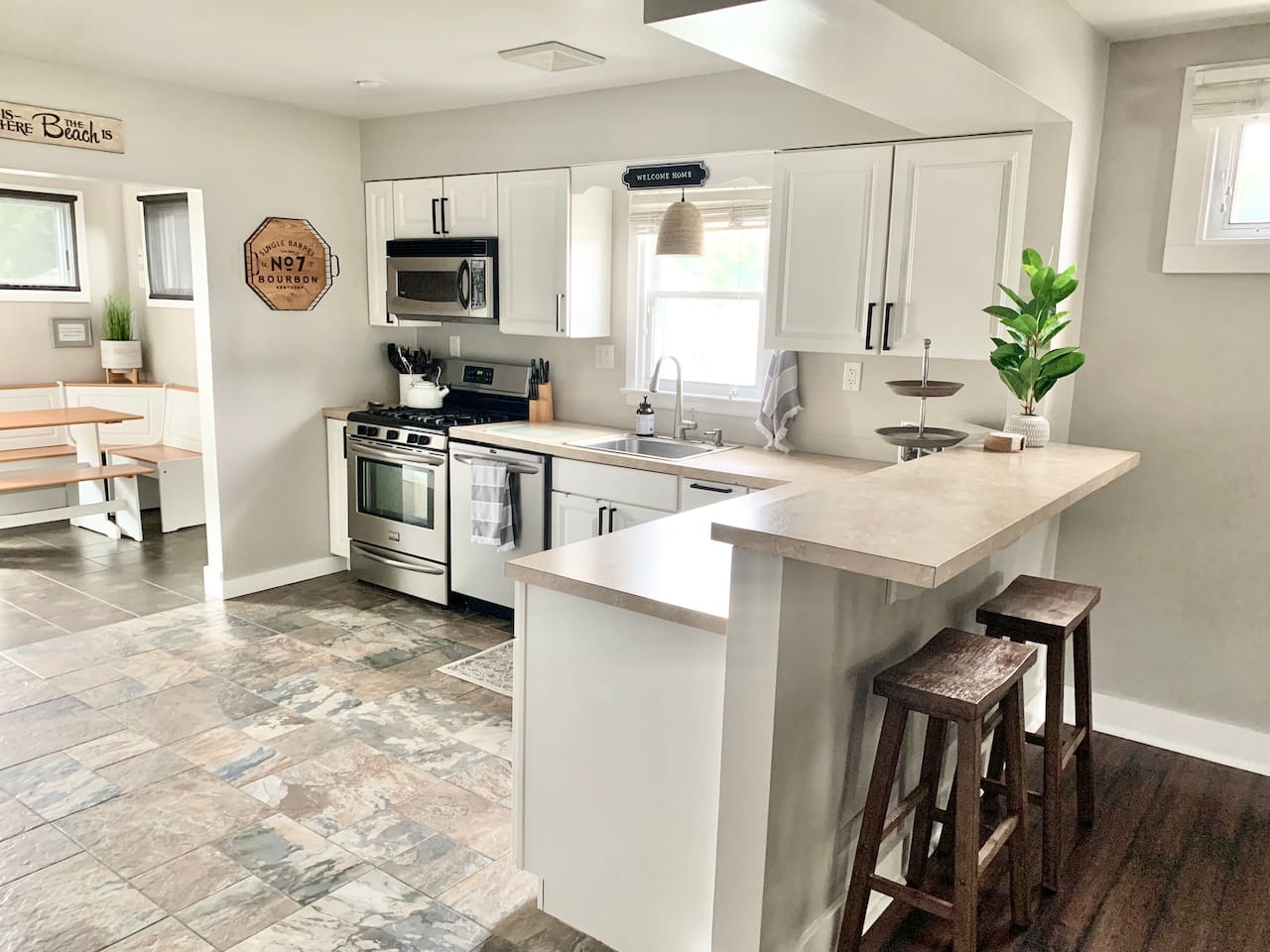 This modern kitchen features stainless steel appliances, a deep sink, and a raised breakfast bar with wooden stools. The open layout leads directly into a sunlit corner breakfast nook, providing a cozy spot for morning coffee.