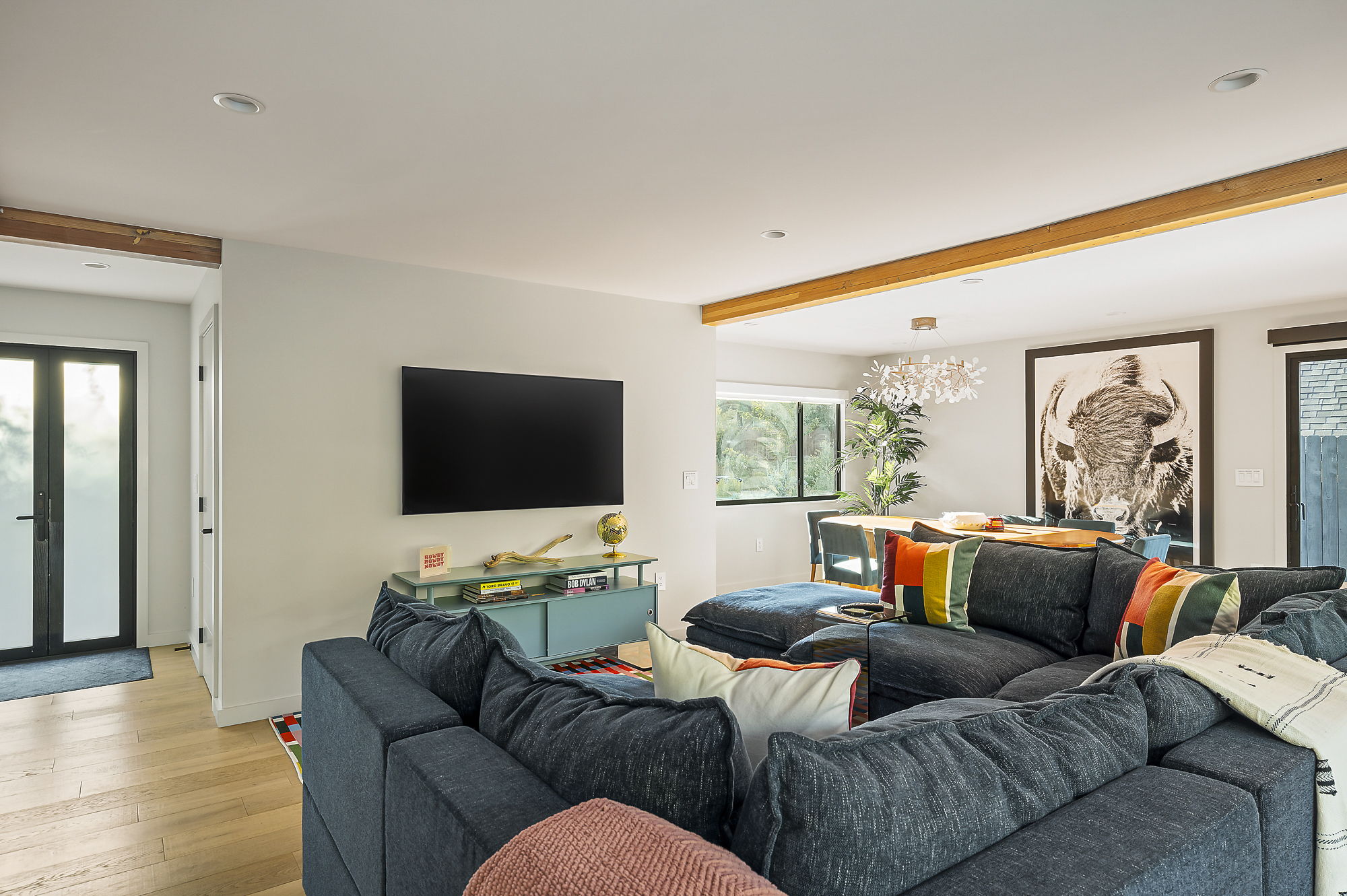 A modern open-concept living area featuring a large sectional sofa, a wall-mounted television, and exposed wooden beams, with a dining area and a large framed buffalo print visible in the background.