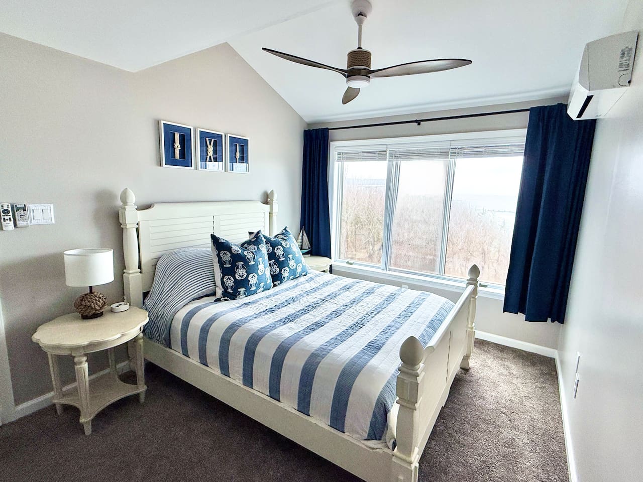 This coastal-themed bedroom features a white bed frame with blue-striped bedding and navy curtains. A bedside table, nautical wall art, and a ceiling fan complete the space with views of the outdoors.