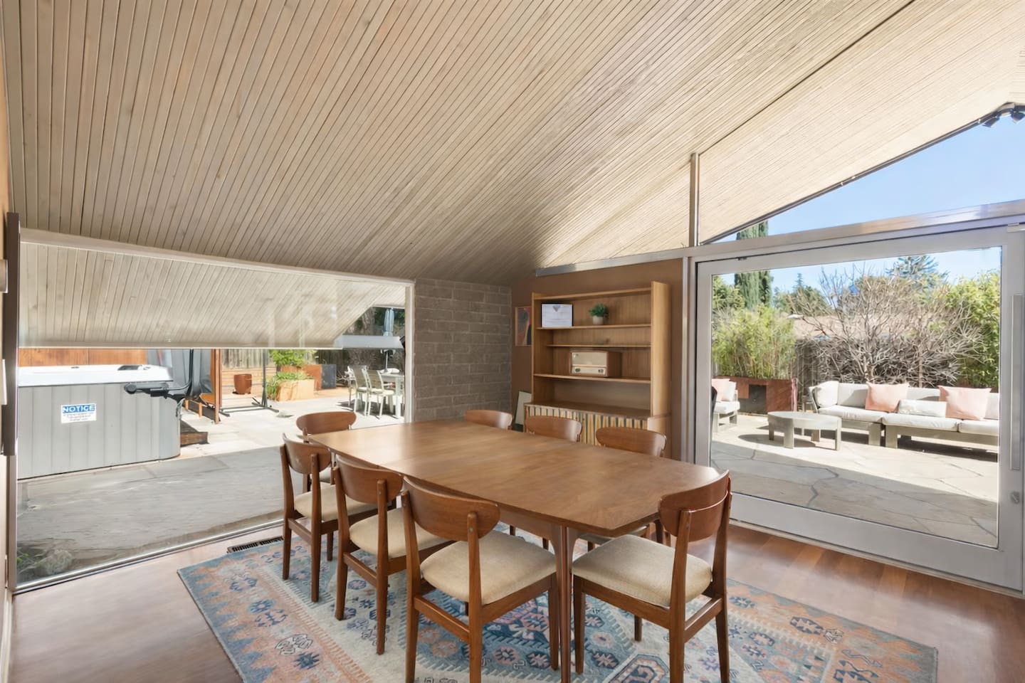 Mid-century dining space with a wooden table for six, vaulted wood ceiling, and floor-to-ceiling glass doors opening to a sunlit patio.