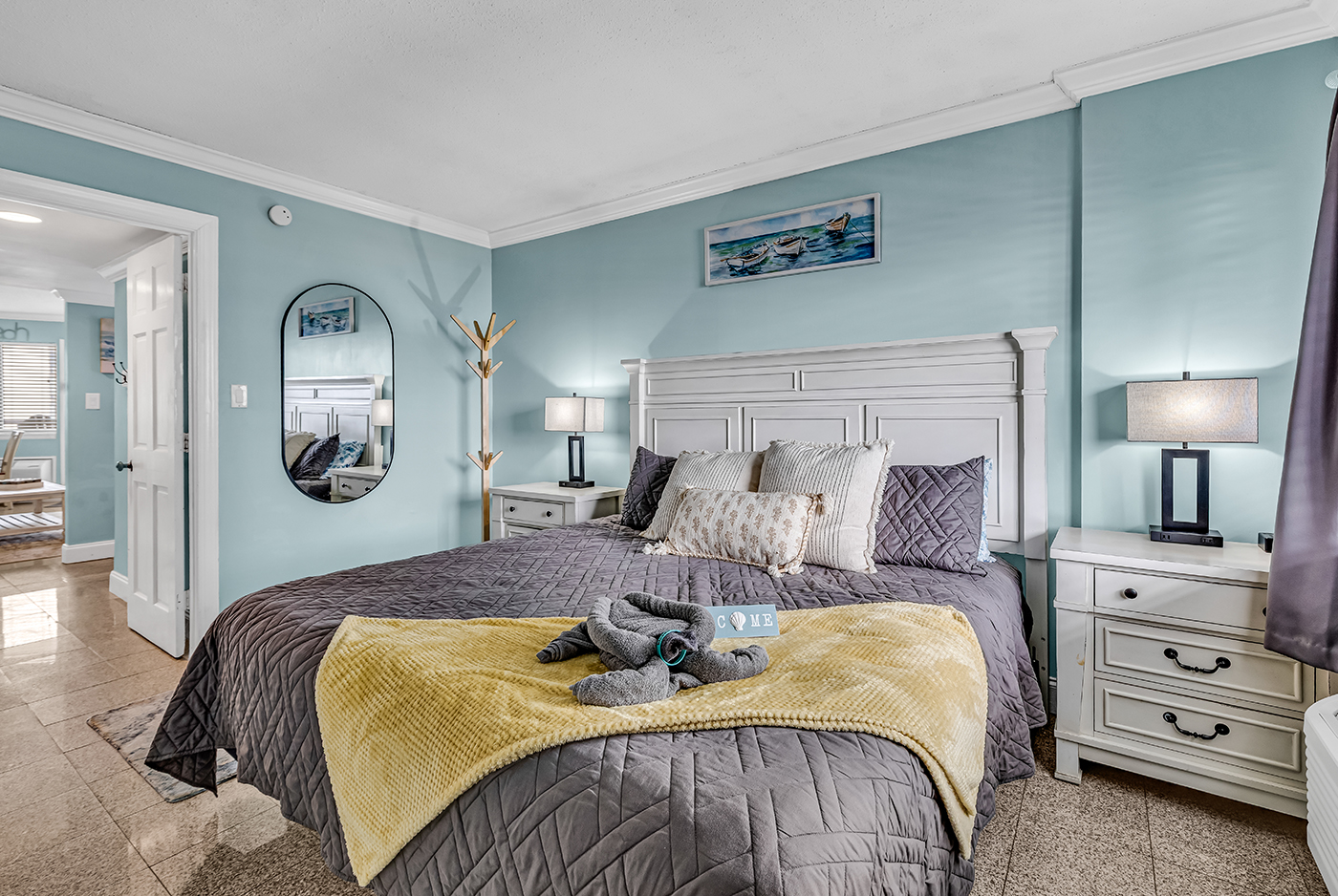 King bedroom with plush bedding, nightstands with lamps, dresser with mirror, and coastal blue décor