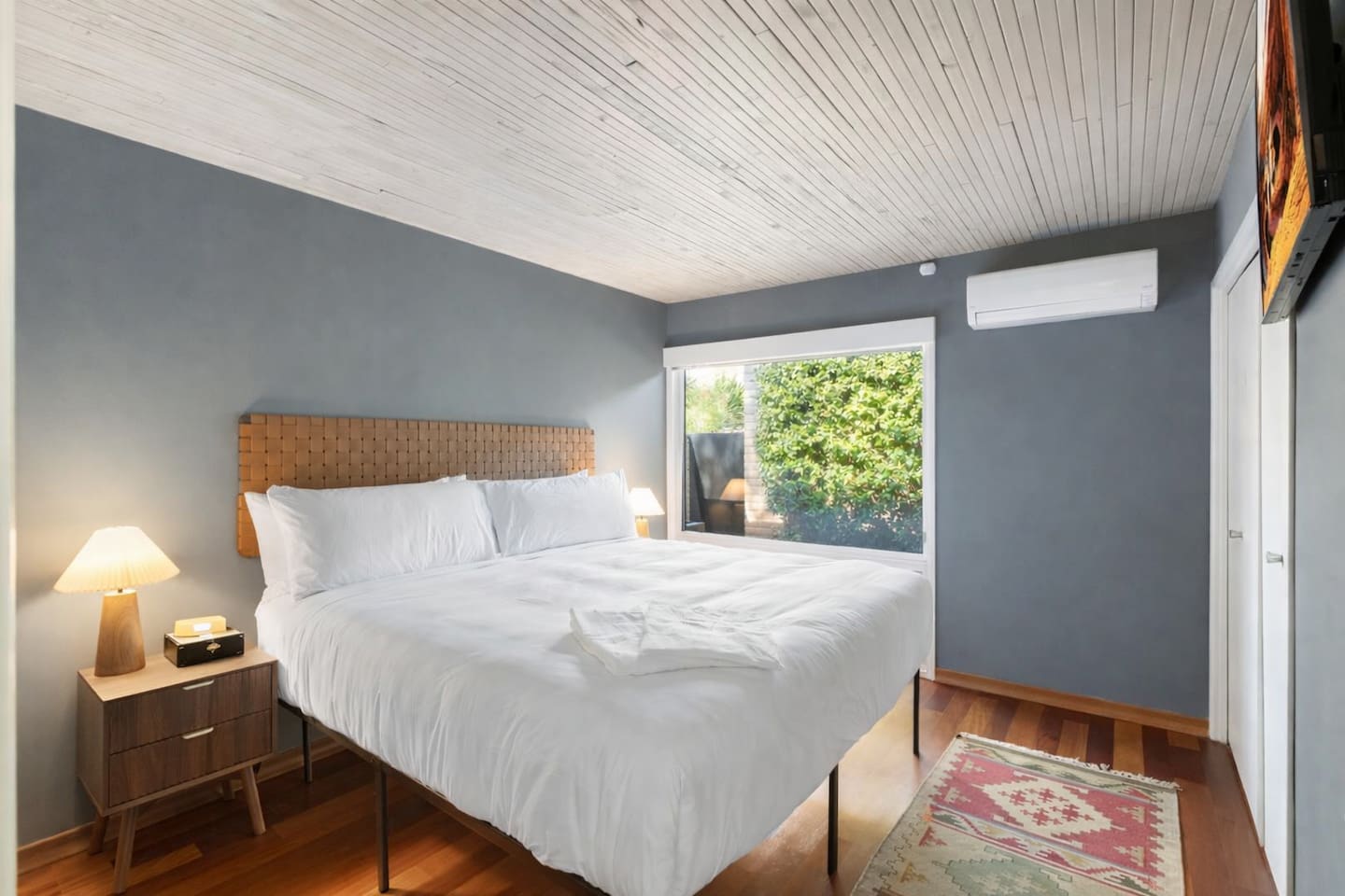 Modern bedroom with a king bed, soft gray walls, warm wood accents, and a large window overlooking greenery.