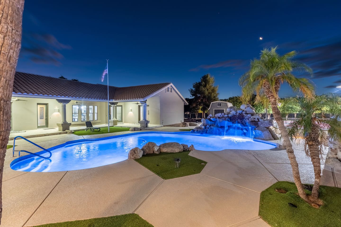 Serene nighttime ambiance featuring a spacious pool with built-in waterfall and lush palm trees.

