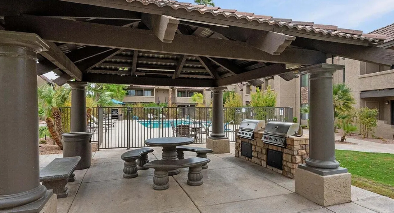Shared outdoor dining area with BBQ grills, shaded seating, and pool area nearby.