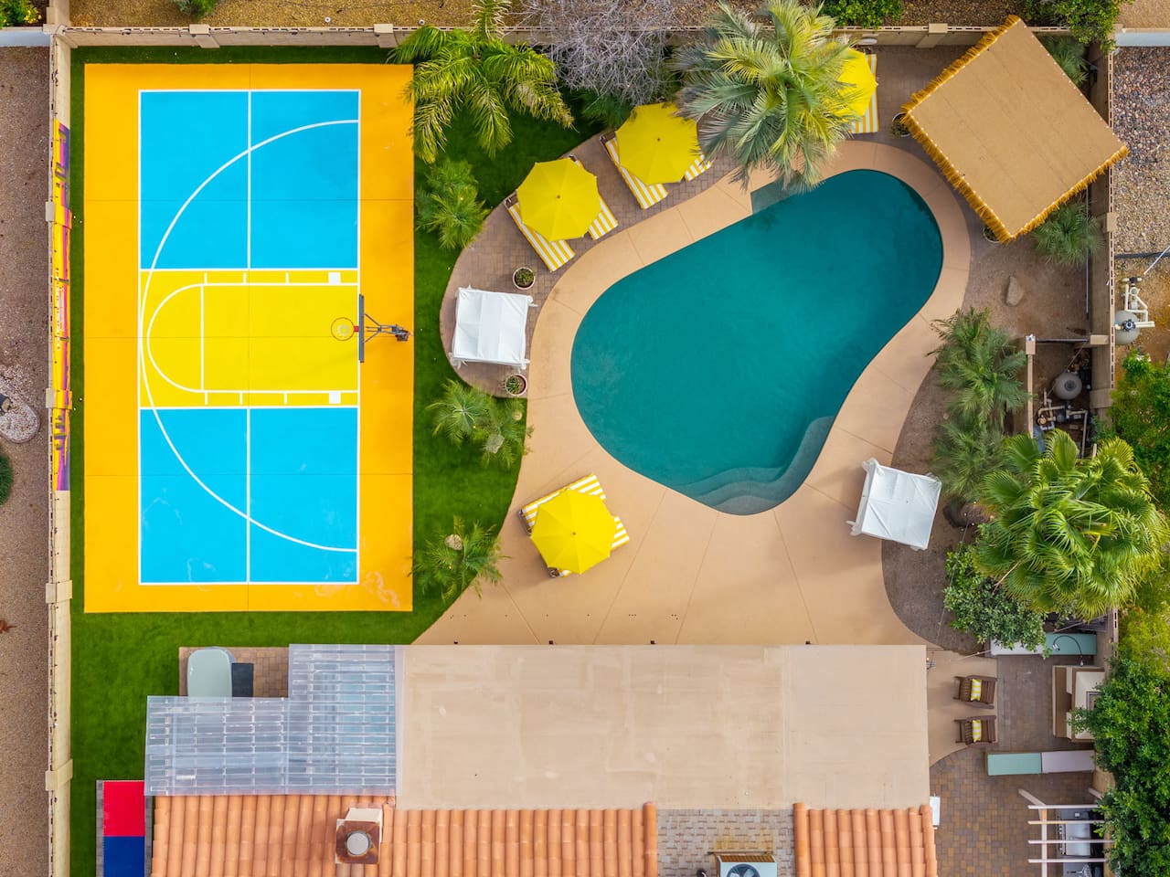 Overhead view of pool and sports court with yellow umbrellas and green landscaping—compact yet complete recreation space for guests.