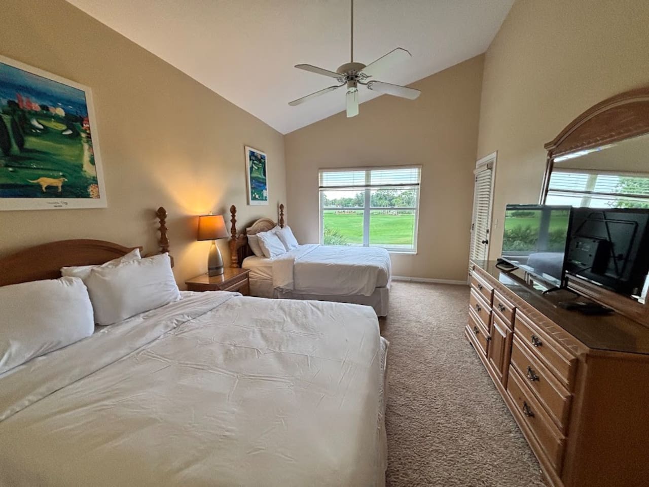 Bright bedroom featuring two beds, TV, dresser storage, ceiling fan, and peaceful green space views.