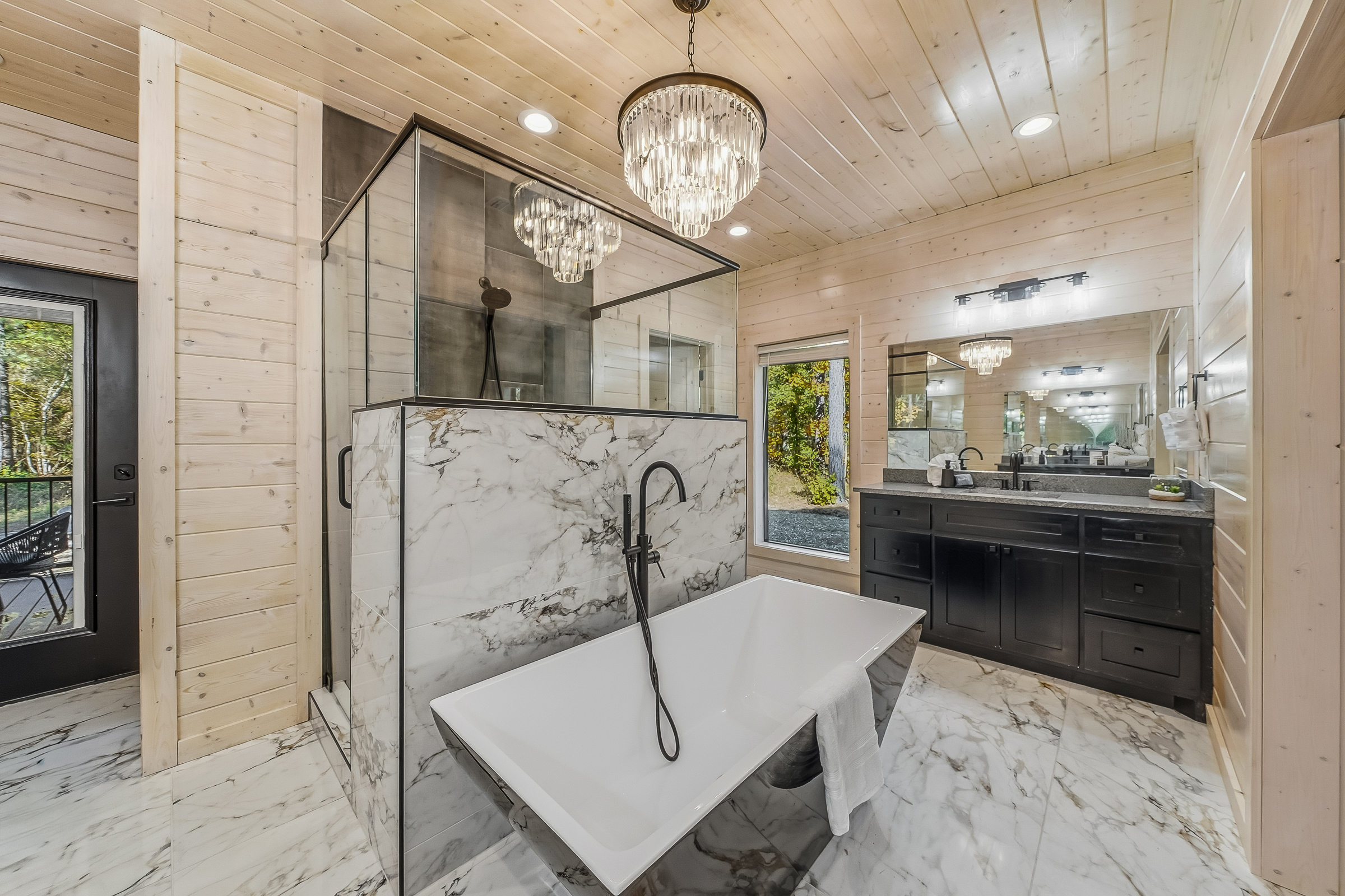 A luxury bathroom centerpiece featuring a sculptural soaking tub under a crystal chandelier and surrounded by forest framed windows.