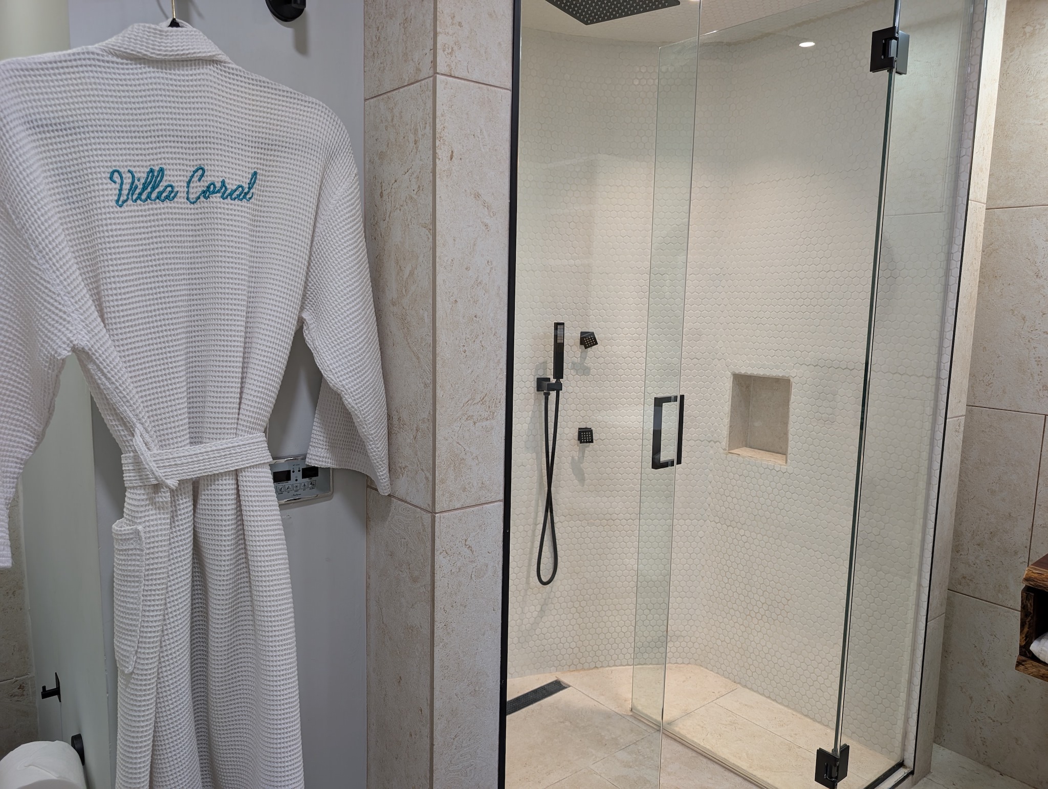 Premium custom-embroidered Villa Coral bath robe hanging next to a glass steam shower enclosure with digital touch control panel in the primary suite bathroom in Dorado Puerto Rico.
