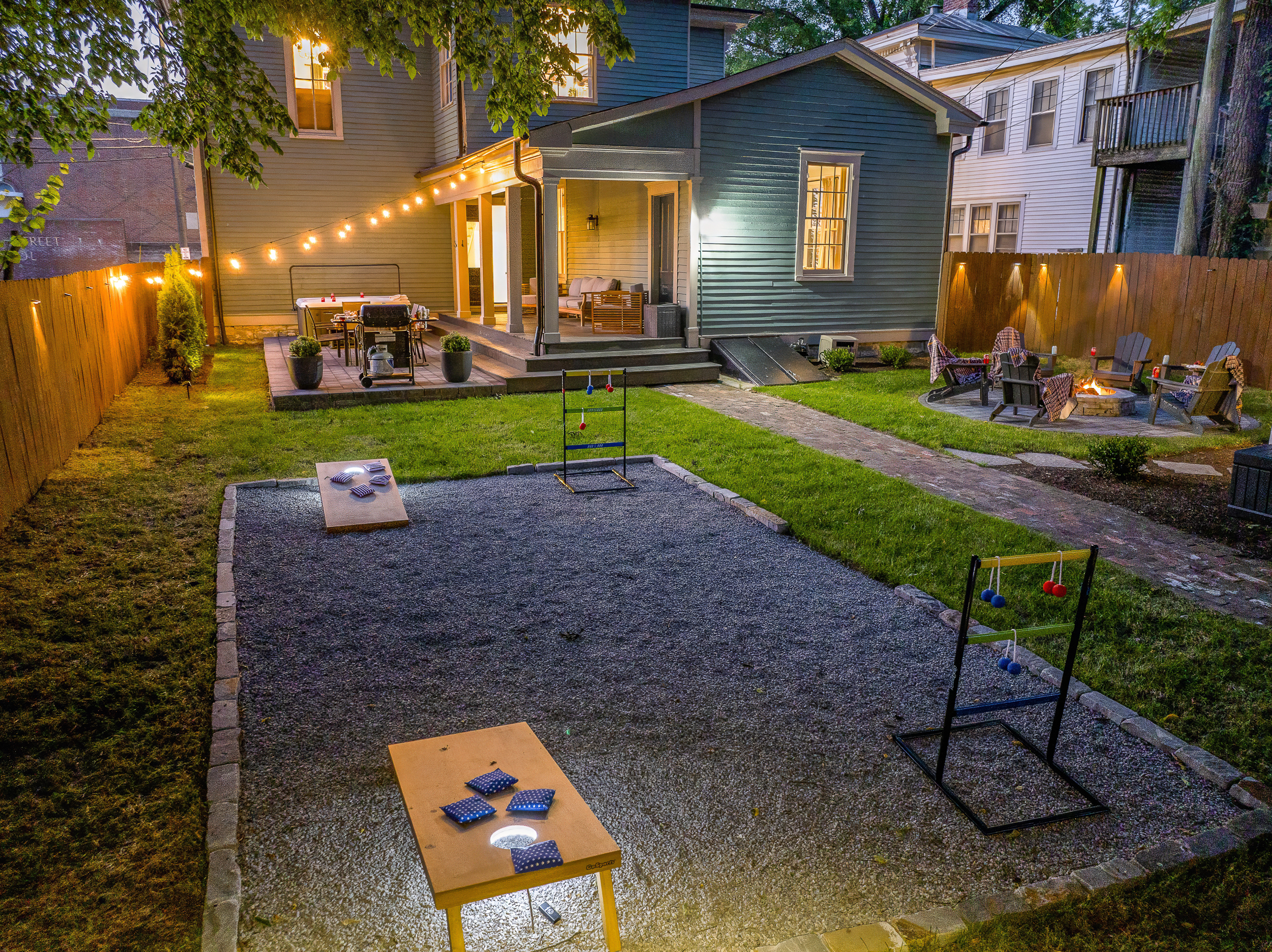 The full backyard at dusk: fire pit lit, game room glowing, covered porch set and ready.